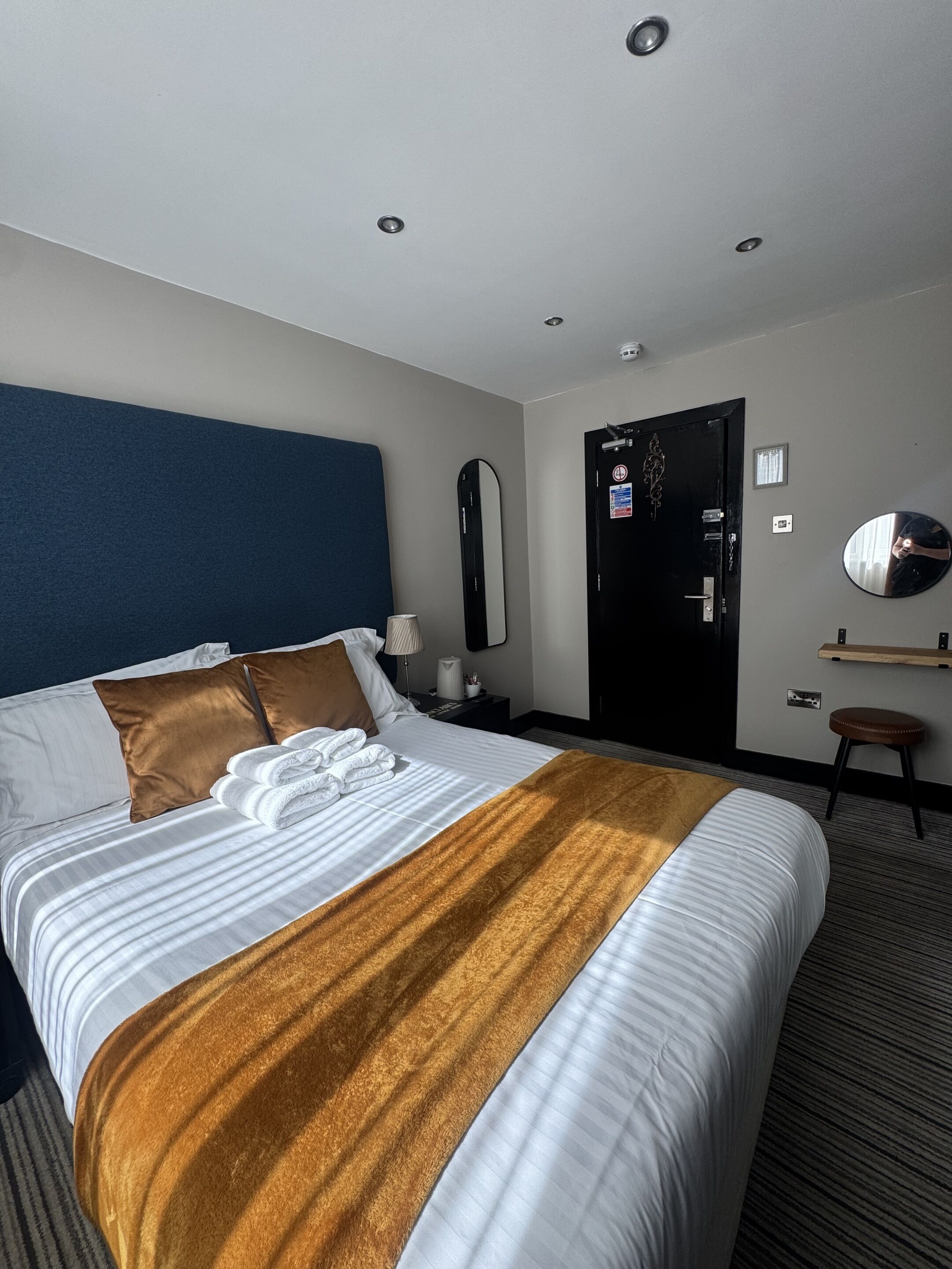 A hotel room with a large bed, a blue headboard, a mirror, a door, and a small stool.