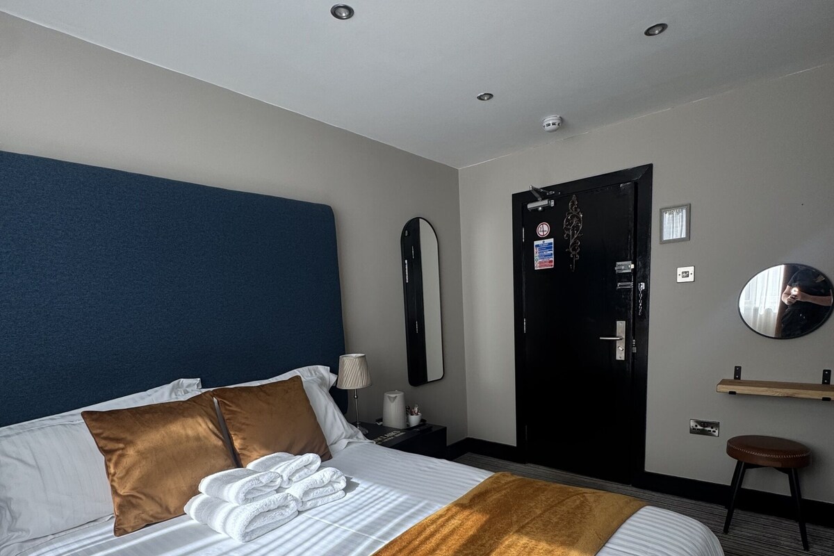 A hotel room with a large bed, a blue headboard, a mirror, a door, and a small stool.