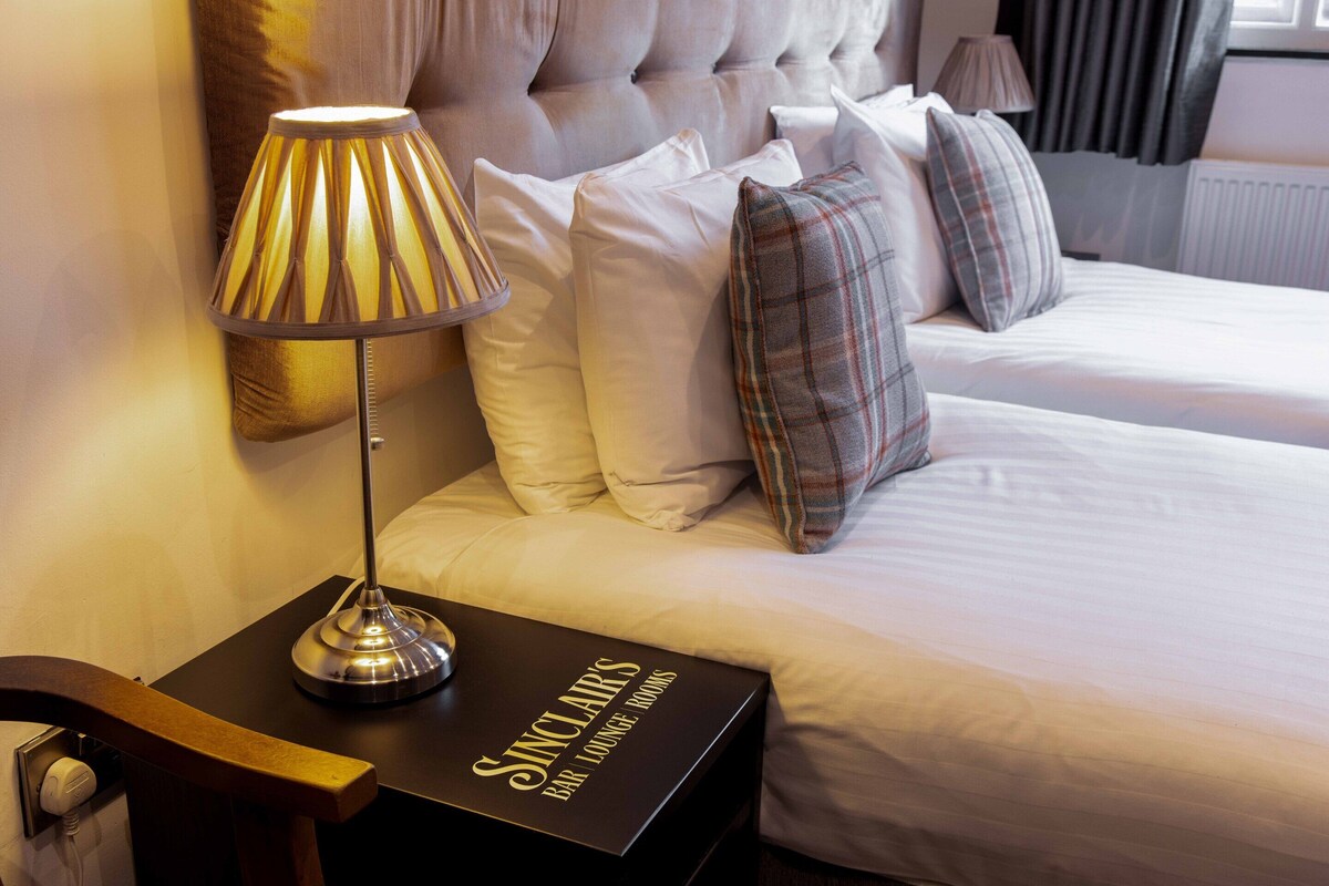 Una habitación de hotel con una cama, una lámpara de noche, una mesita de noche y una caja con la etiqueta 'Sinclair's Bar Lounge Rooms'.