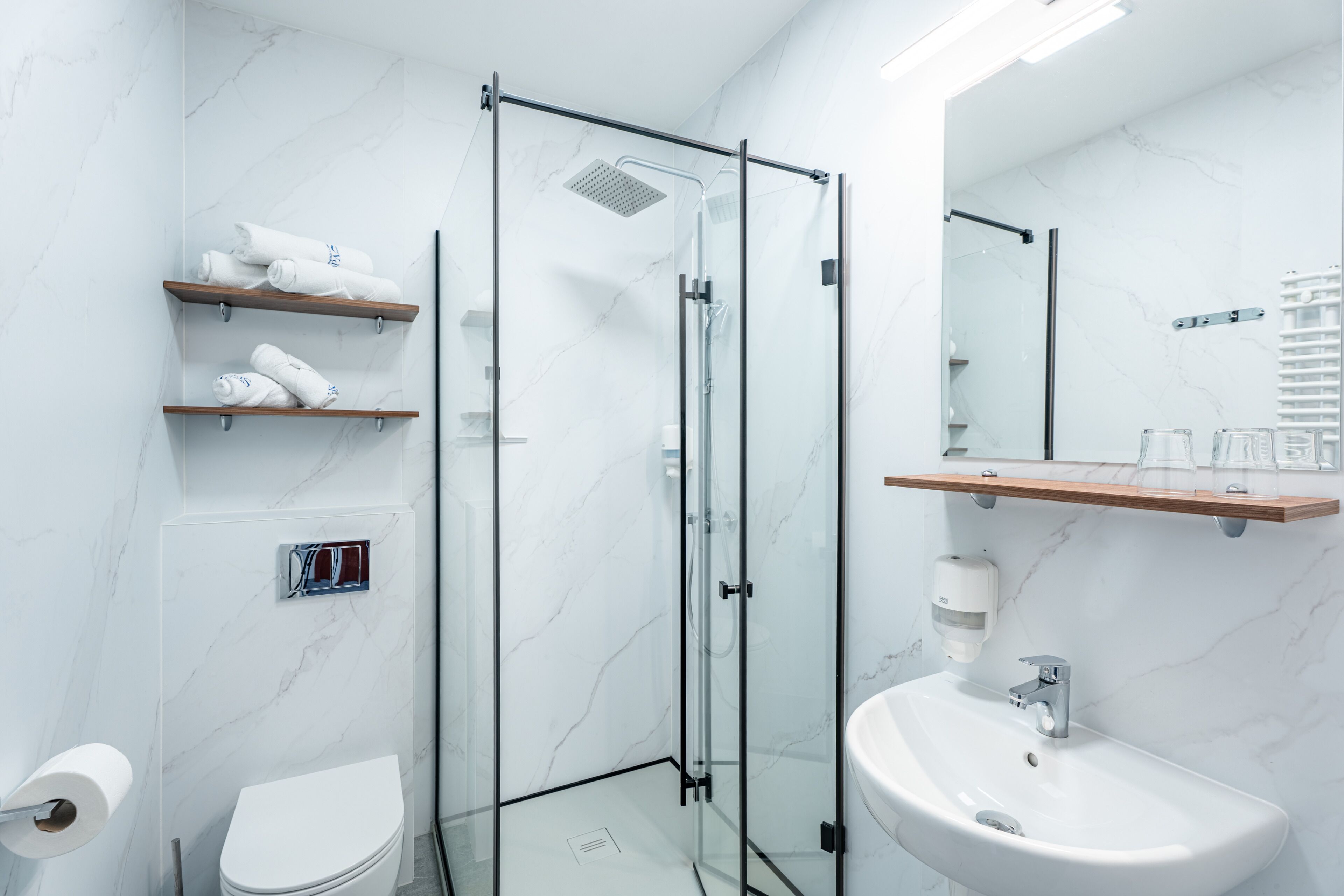 A modern bathroom with a glass-enclosed shower, a white sink, and a toilet. There are wooden shelves with towels and a soap dispenser.