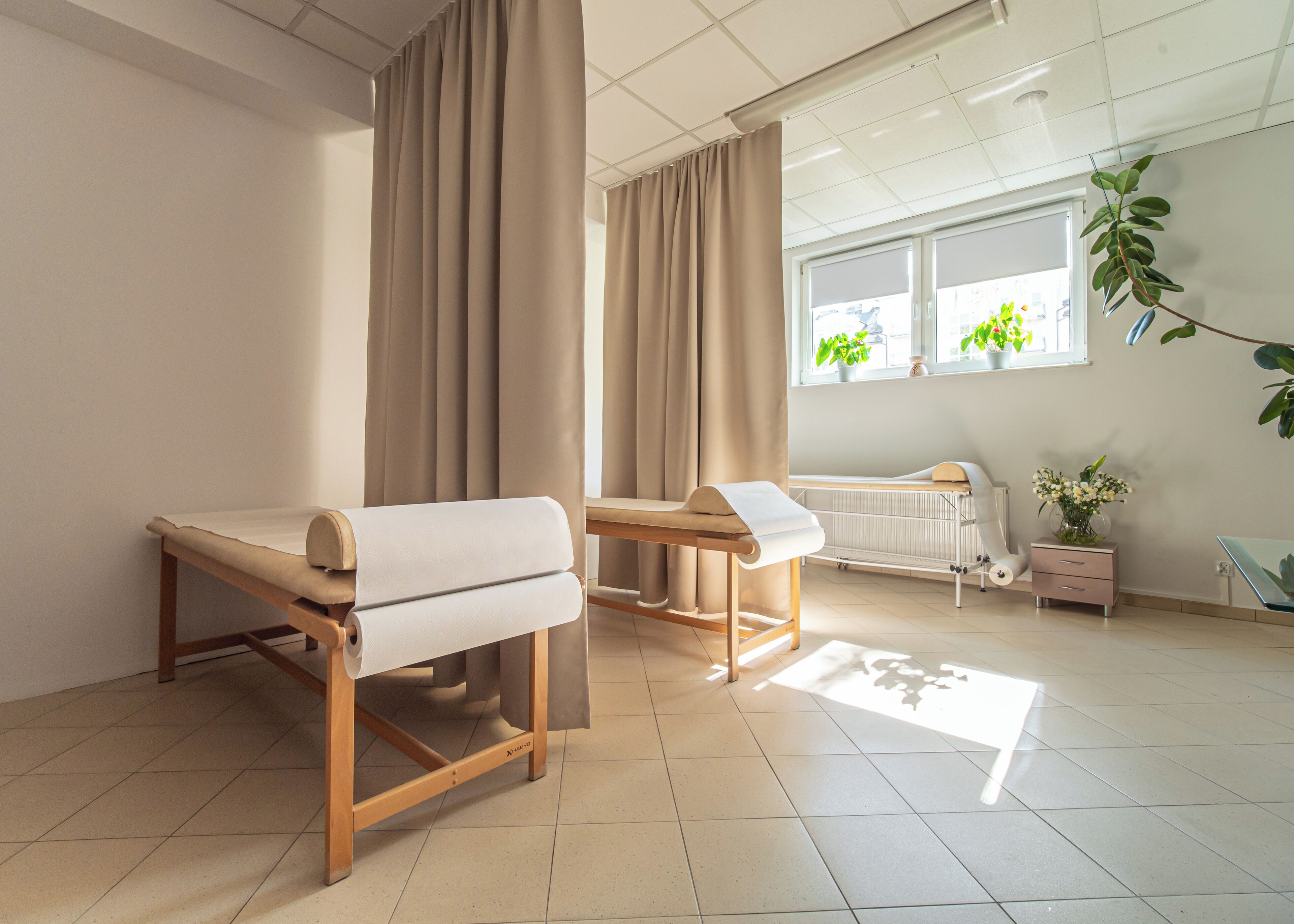Una habitación limpia y bien iluminada con una camilla de madera, una ventana con plantas y un radiador.