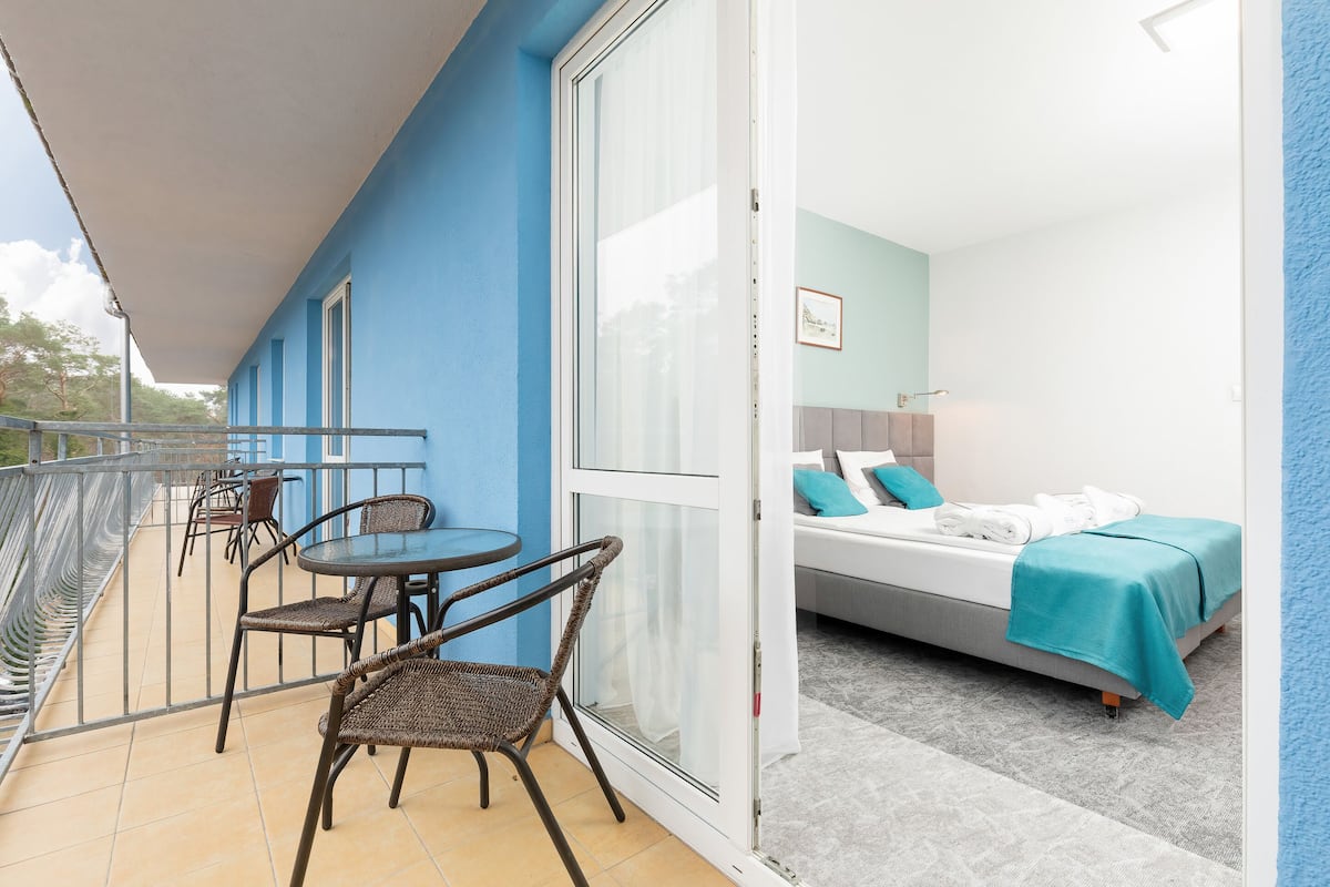 Un balcón con sillas de mimbre y una mesa, una cama con colcha color turquesa, y vista al cielo.