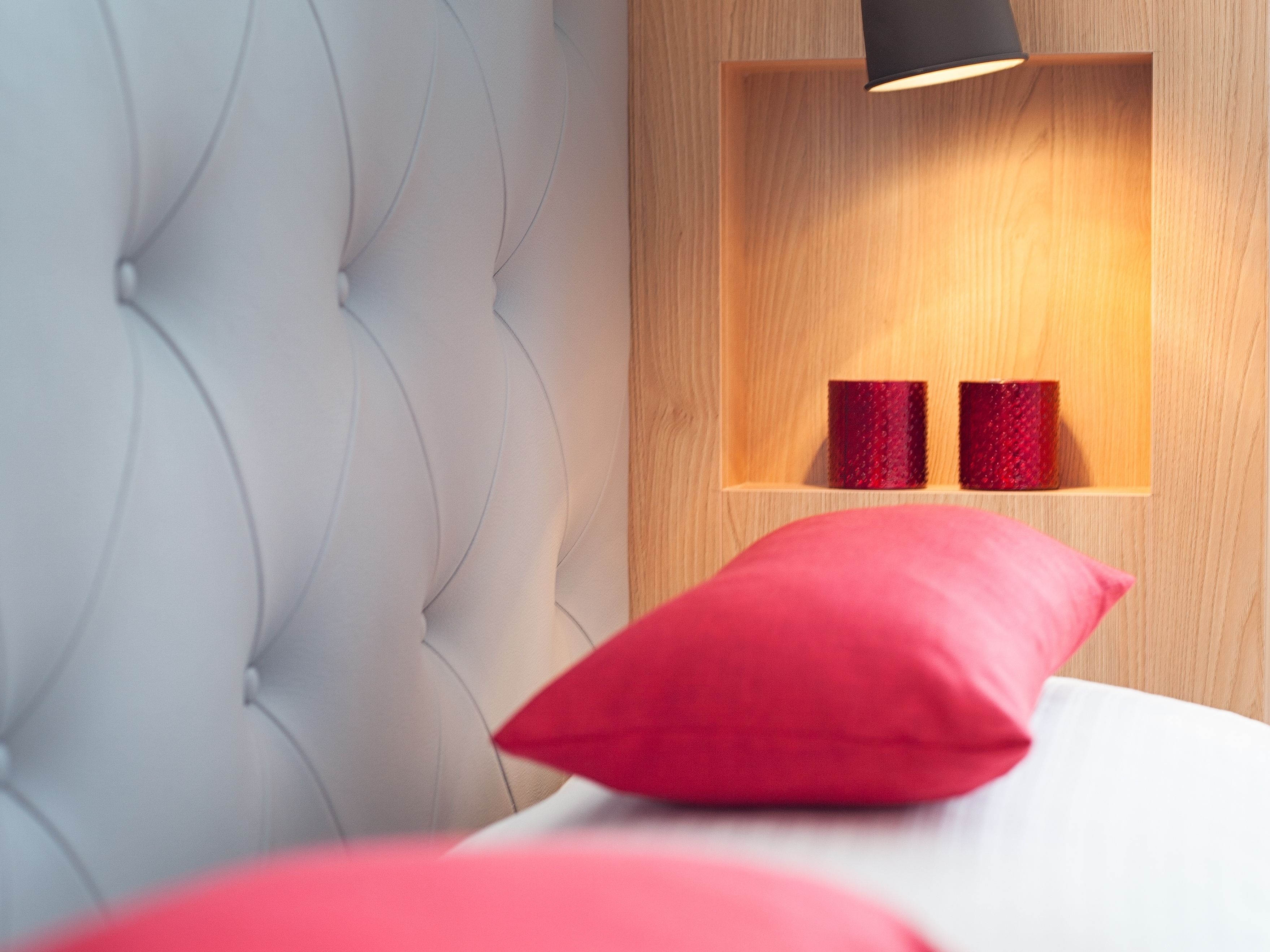 A bedroom with a tufted headboard, a wooden bedside table with two red decorative items, and a red pillow on the bed.