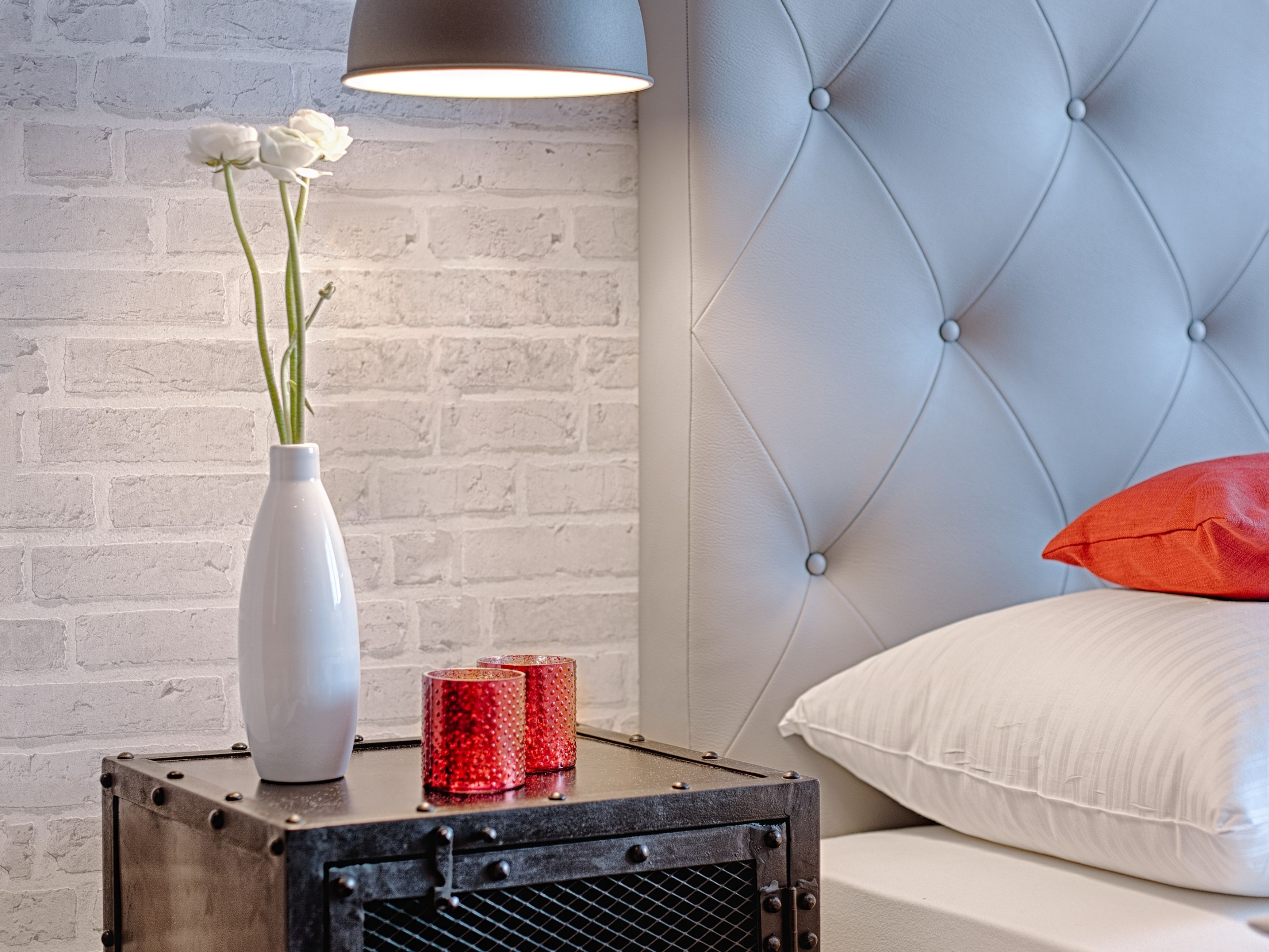 Eine weiße Ziegelwand, ein gestepptes Kopfteil, ein weißes Kissen, ein rotes Kissen, eine weiße Vase mit Blumen und zwei glitzernde rote Kerzen auf einem Metalltisch.