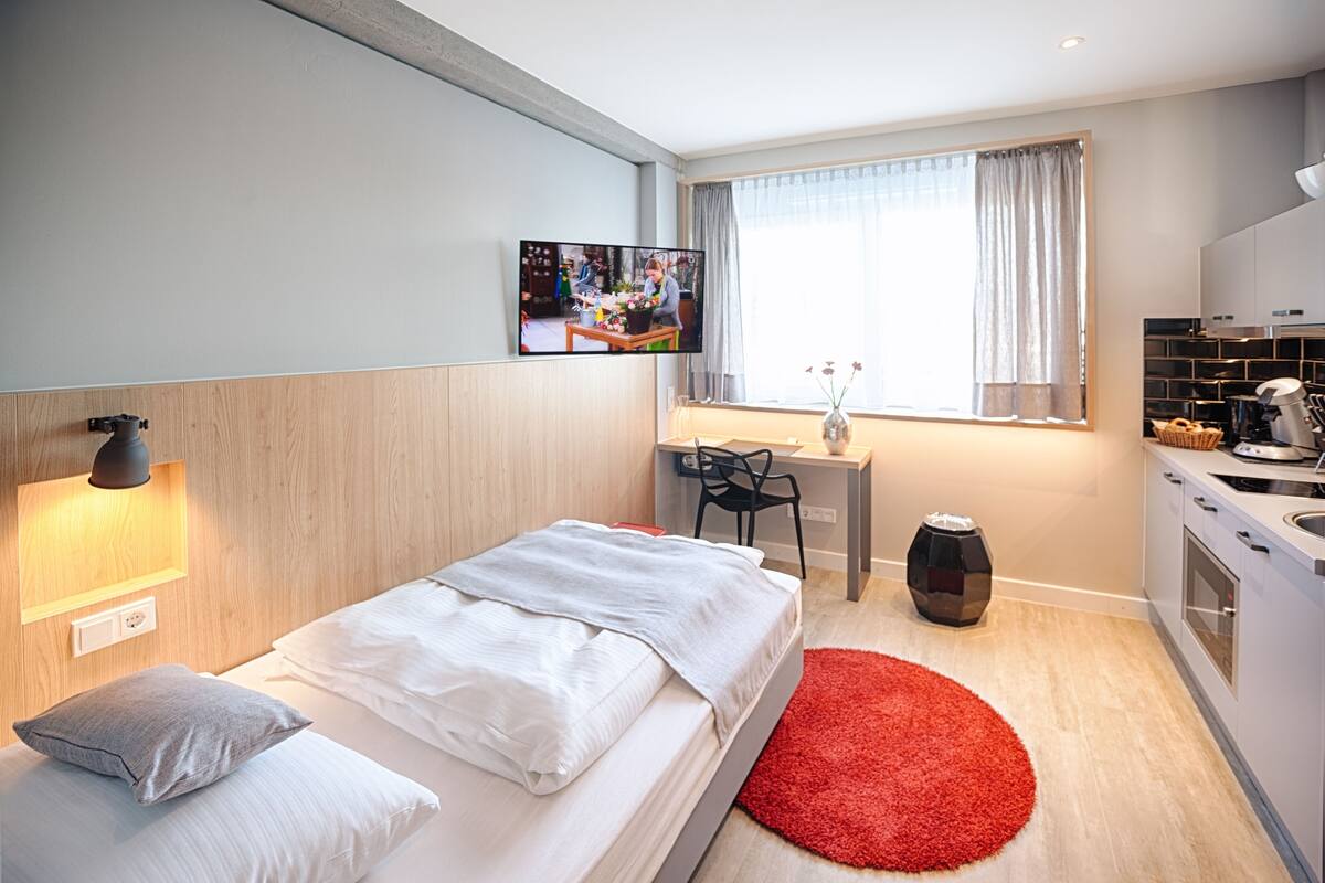 A modern hotel room with a bed, a desk, a chair, a TV, and a kitchen area.