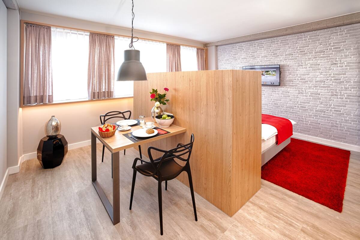 A compact hotel room with a dining area, a bed with a red accent, a brick-patterned wall, and a wooden partition.