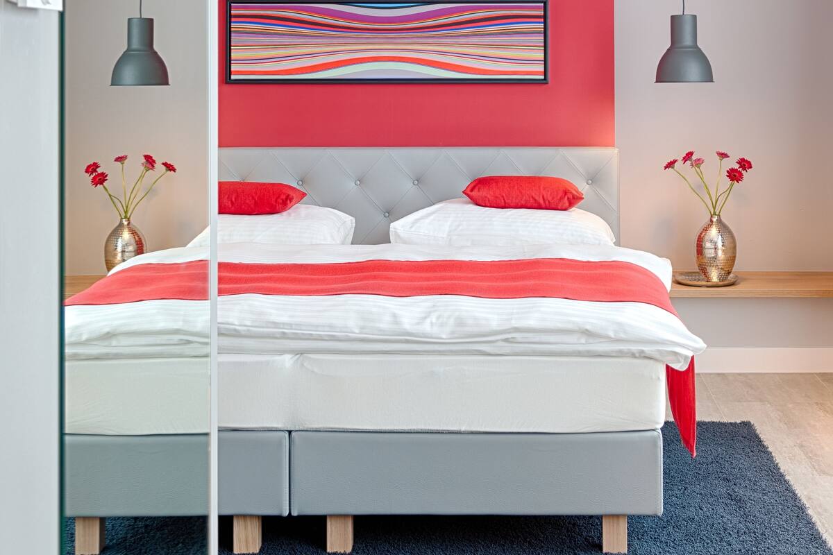 A modern bedroom with a bed, red pillows, a colorful abstract painting, and two vases with red flowers.