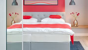 A modern bedroom with a bed, red pillows, a colorful abstract painting, and two vases with red flowers.