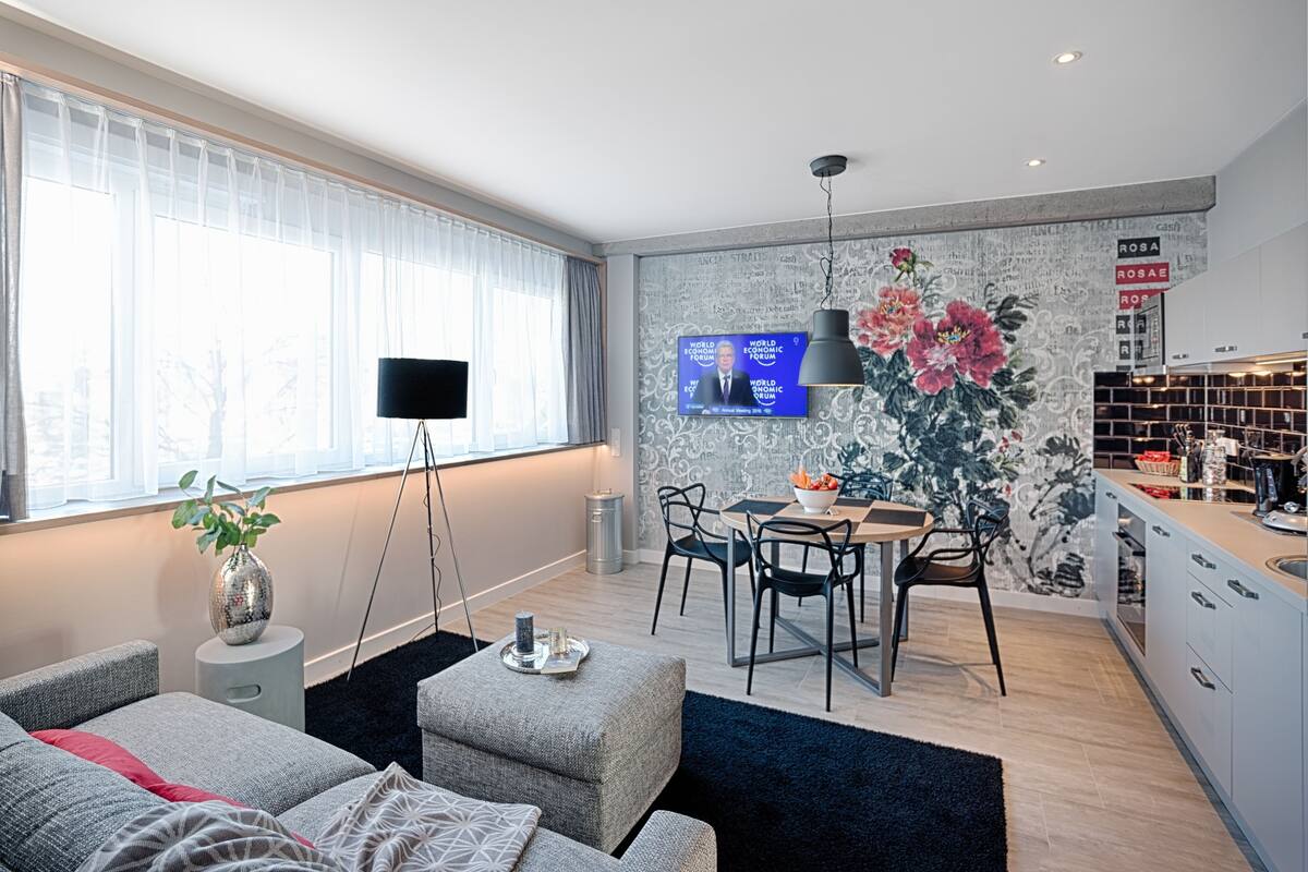 A modern living room with a sofa, a coffee table, and a TV mounted on the wall. There is a dining area with a table and chairs, and a kitchen area with cabinets and a countertop.