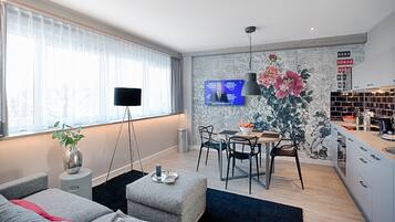 A modern living room with a sofa, a coffee table, and a TV mounted on the wall. There is a dining area with a table and chairs, and a kitchen area with cabinets and a countertop.