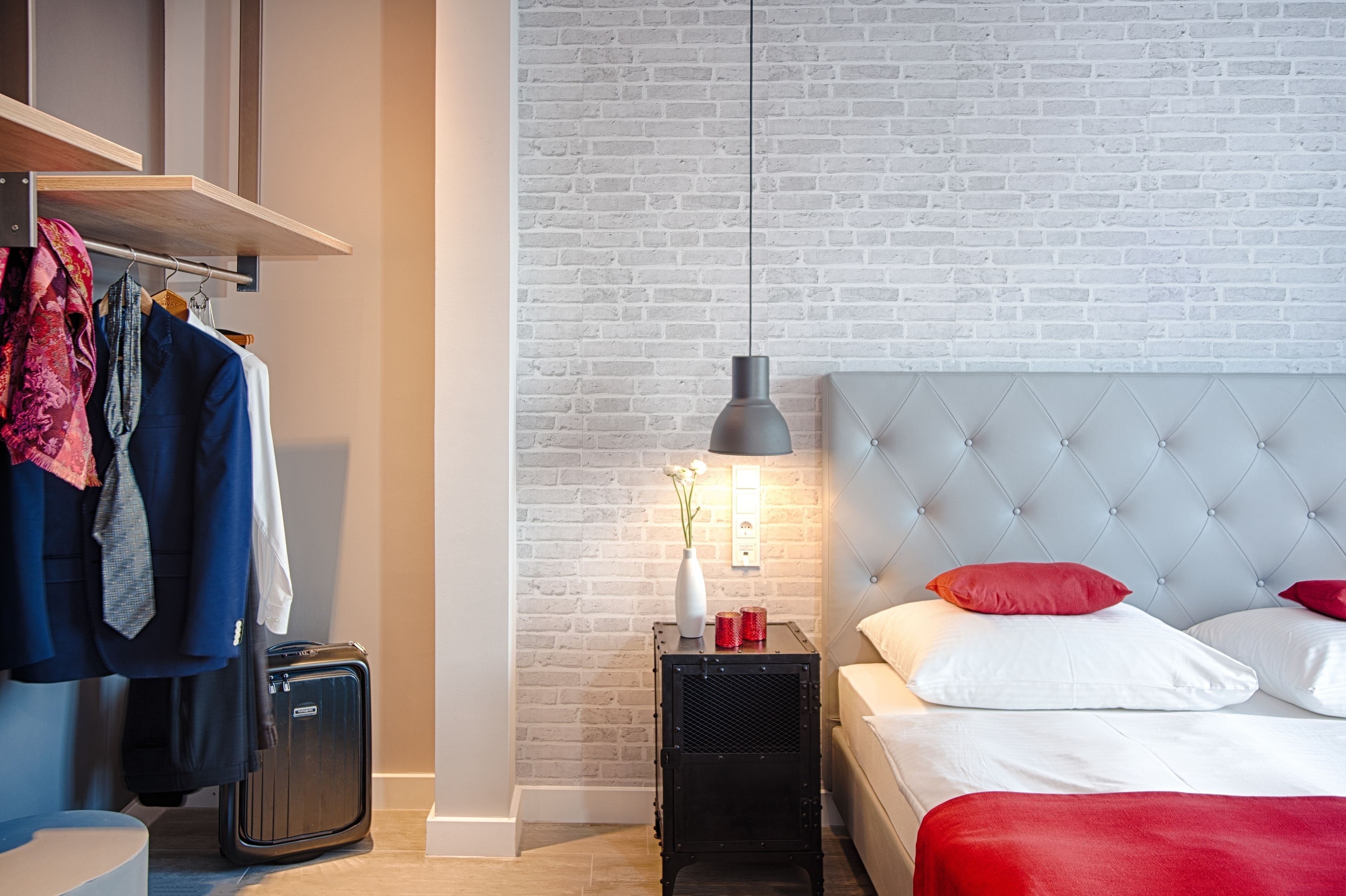 A hotel room with a white brick wall, a tufted headboard, a bed with red and white pillows, a bedside table with a lamp, and a wardrobe with hanging clothes.