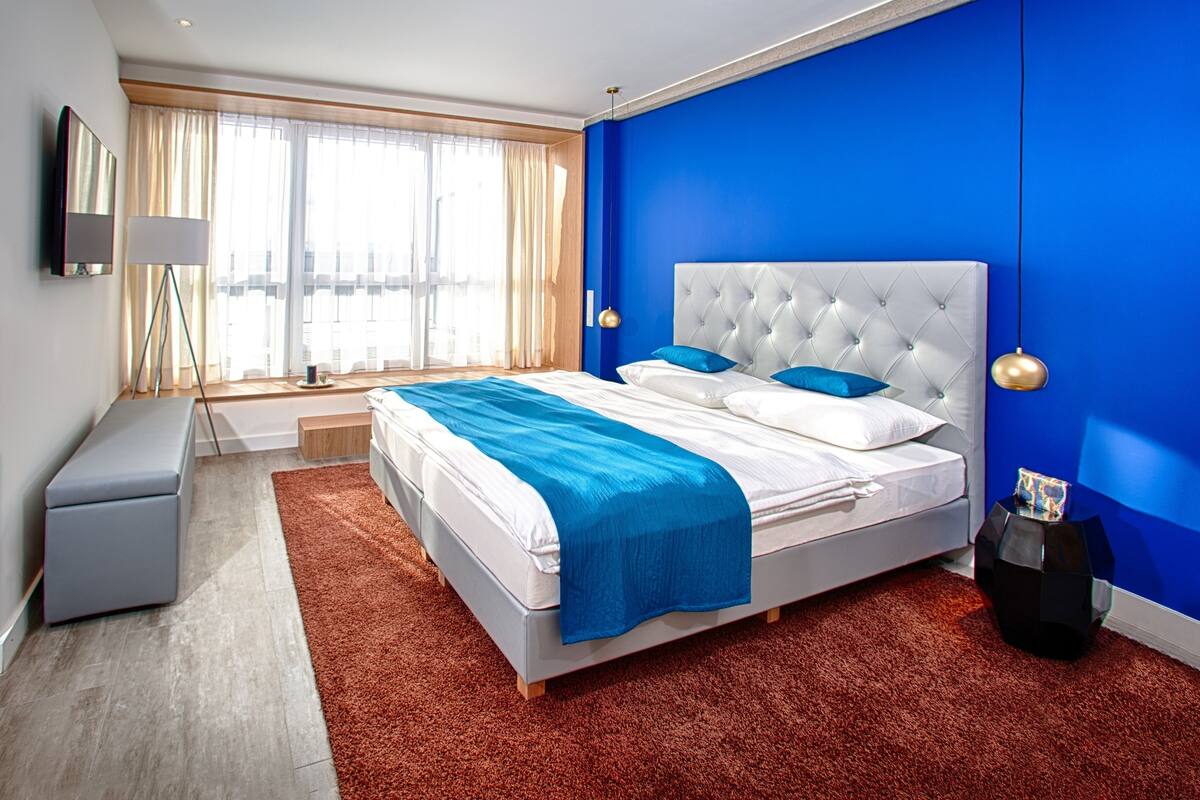 A modern bedroom with a large bed, a bedside table, a lamp, and a view of the city through large windows.