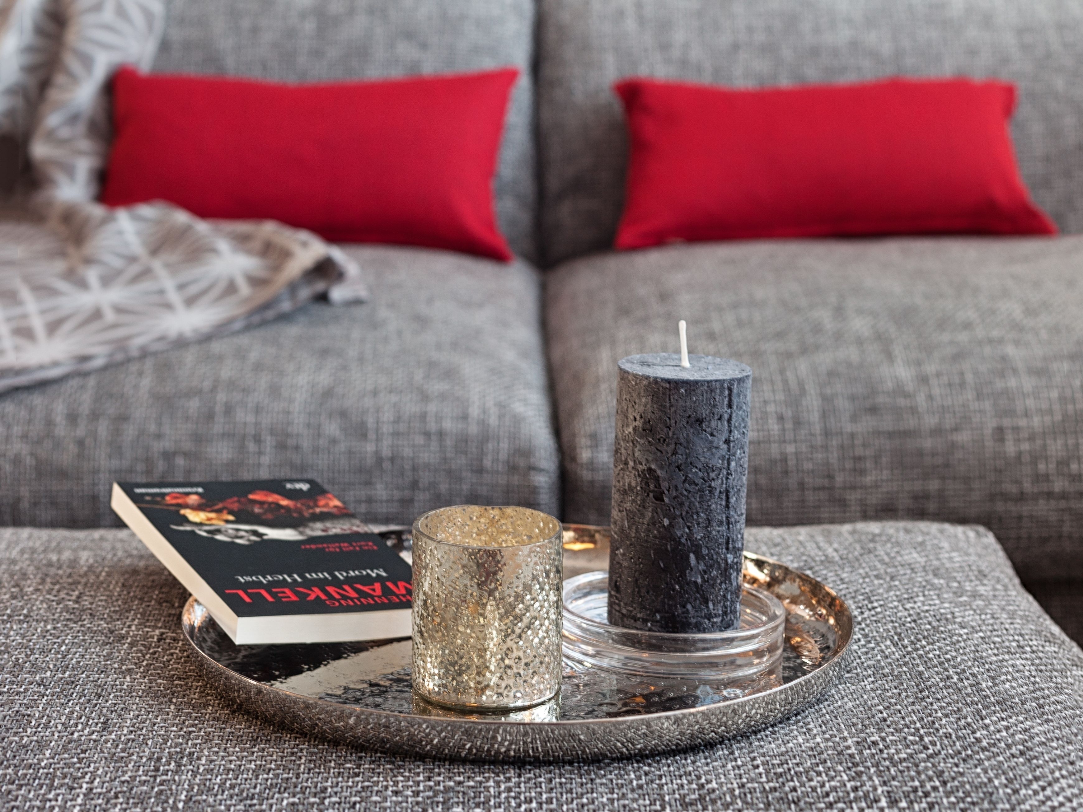 Ein Tablett mit einem Buch, einer Kerze und einem dekorativen Gegenstand auf einem strukturierten Sofa mit roten Kissen.