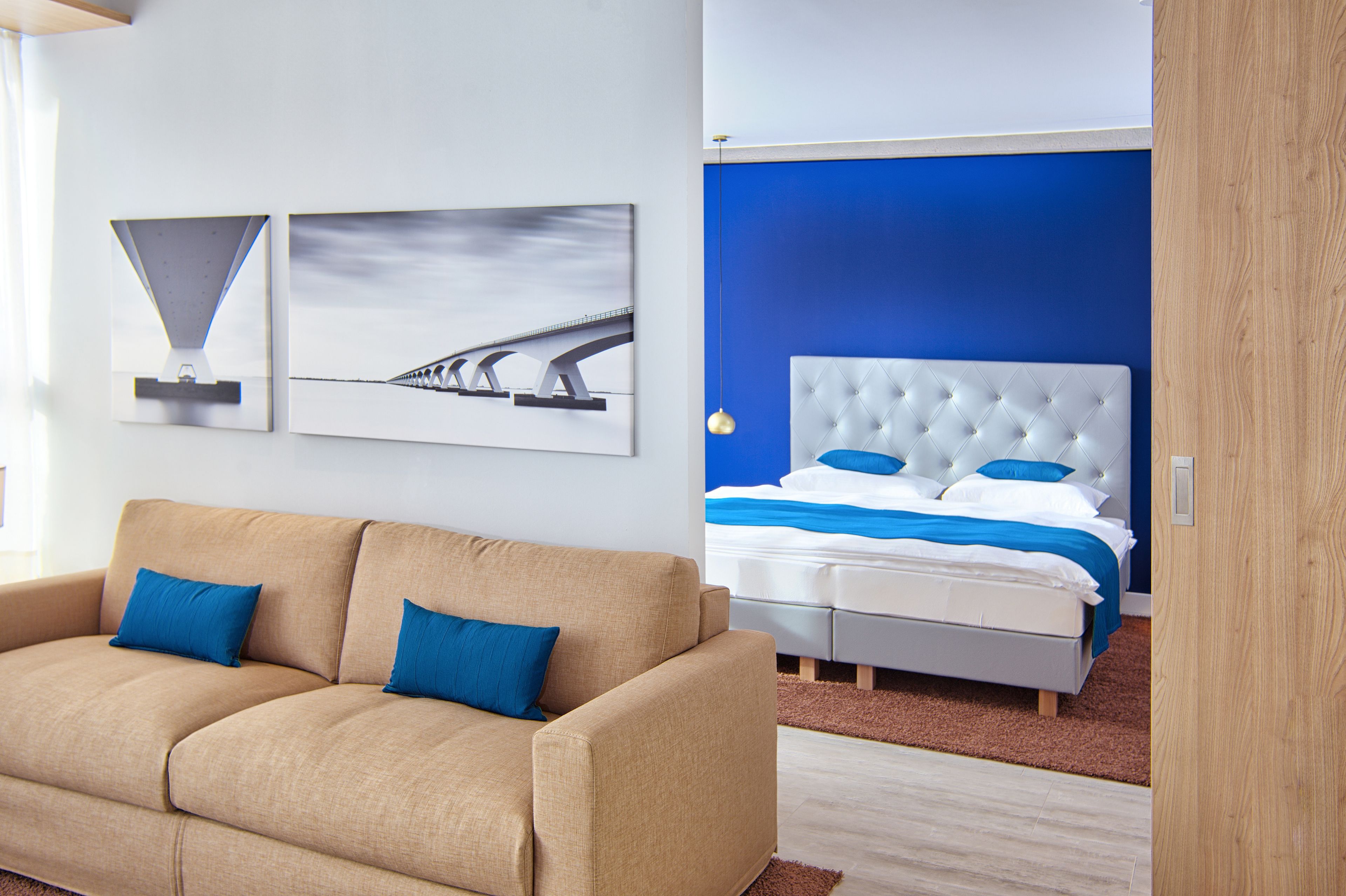 Une chambre d’hôtel moderne avec un canapé beige, un grand lit doté d’une tête de lit capitonnée et deux tableaux encadrés accrochés au mur.