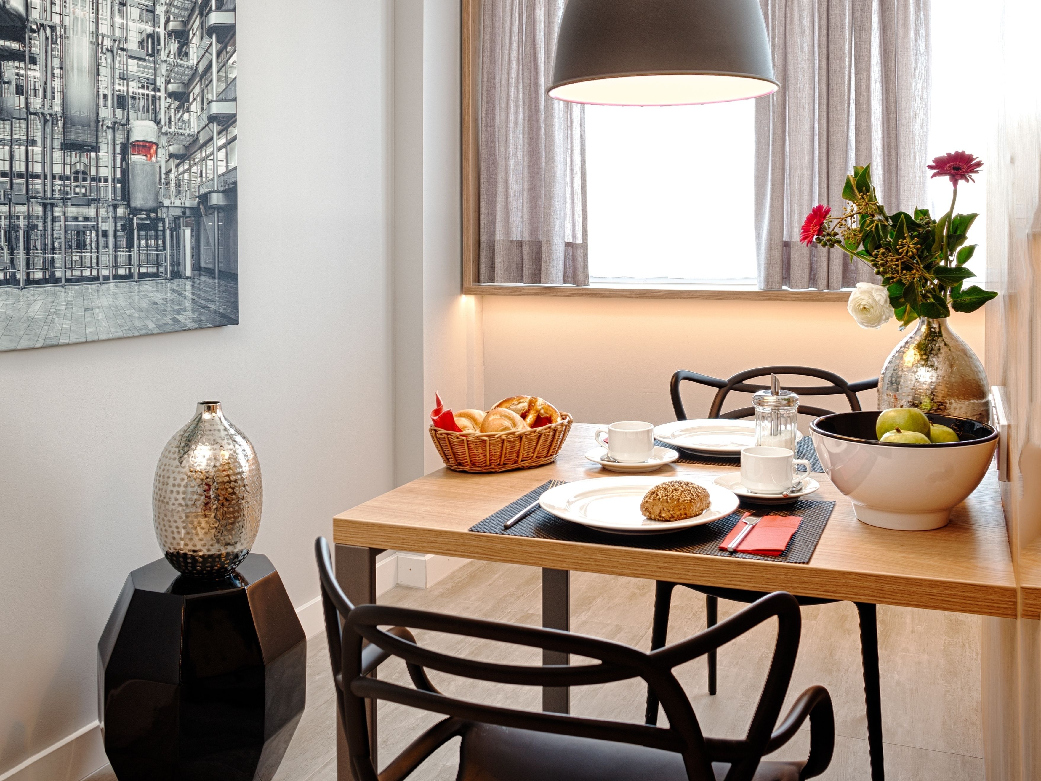 Een eethoek met een houten tafel, stoelen, een vaas met bloemen, een broodmand en een schilderij aan de muur.