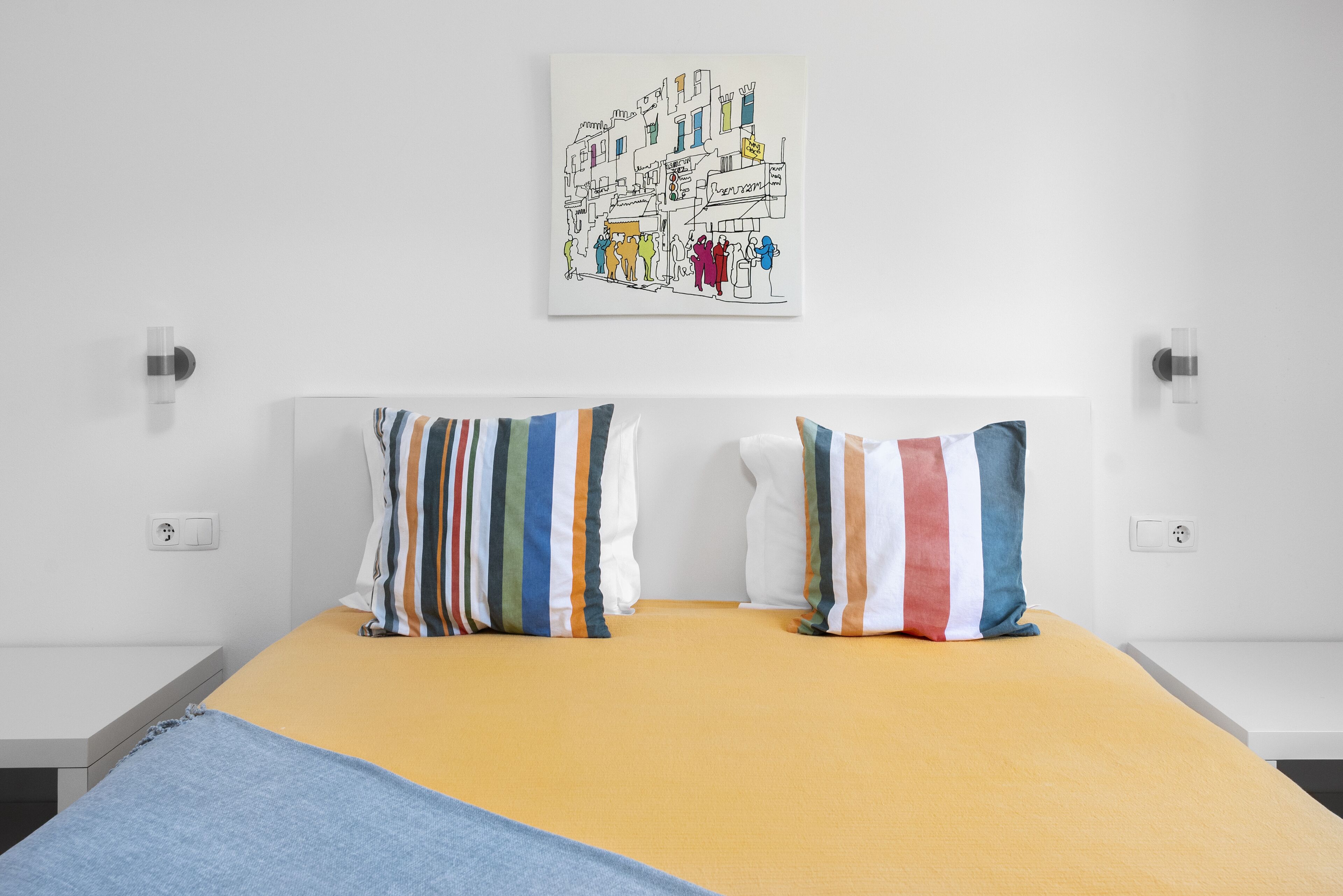 Un lit avec un matelas jaune, deux oreillers à rayures et une œuvre d’art accrochée au mur représentant une scène de rue.