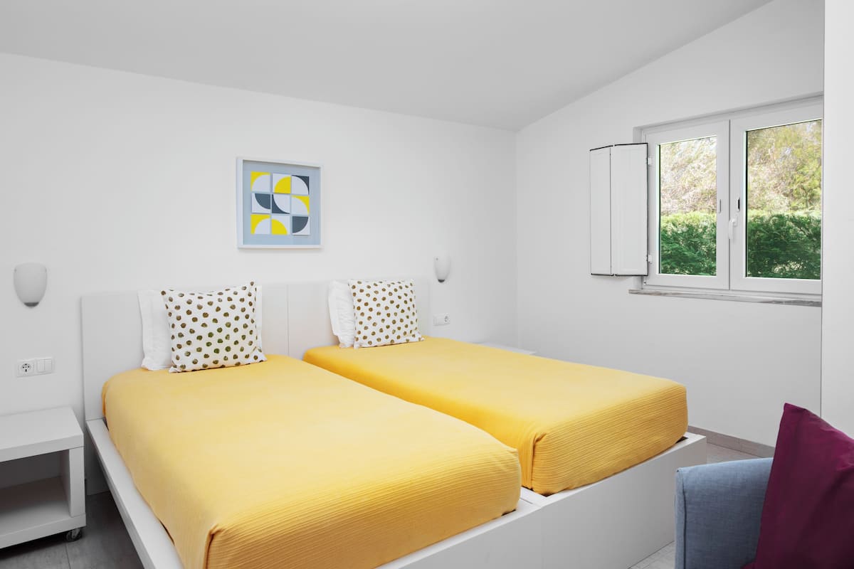 A modern hotel room with two single beds, a window with a view of greenery, a white wall with a geometric art piece, and a small desk with a lamp.