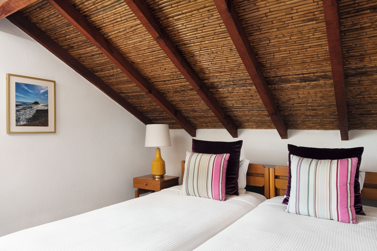 A bedroom with a wooden ceiling, two beds with striped pillows, a nightstand with a lamp, and a framed picture on the wall.