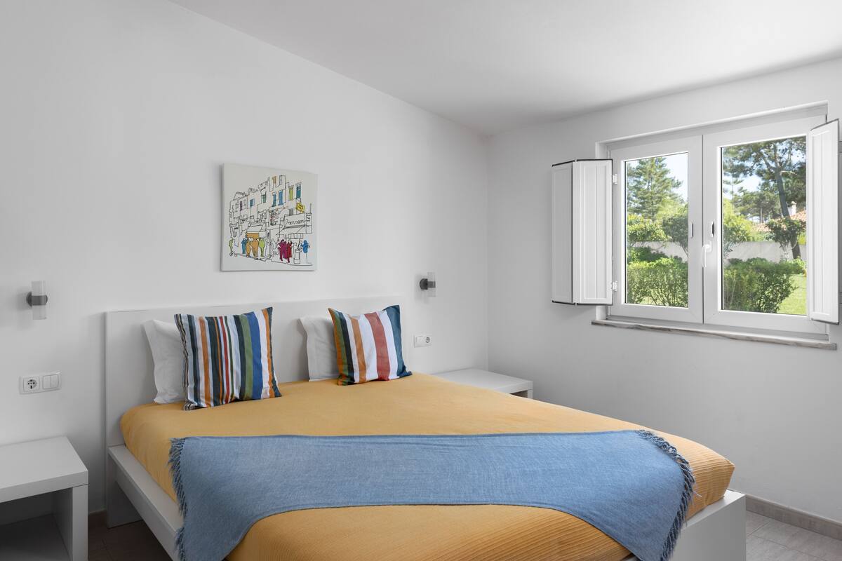 A bedroom with a bed, white nightstands, a window with open shutters, and a wall-mounted artwork.