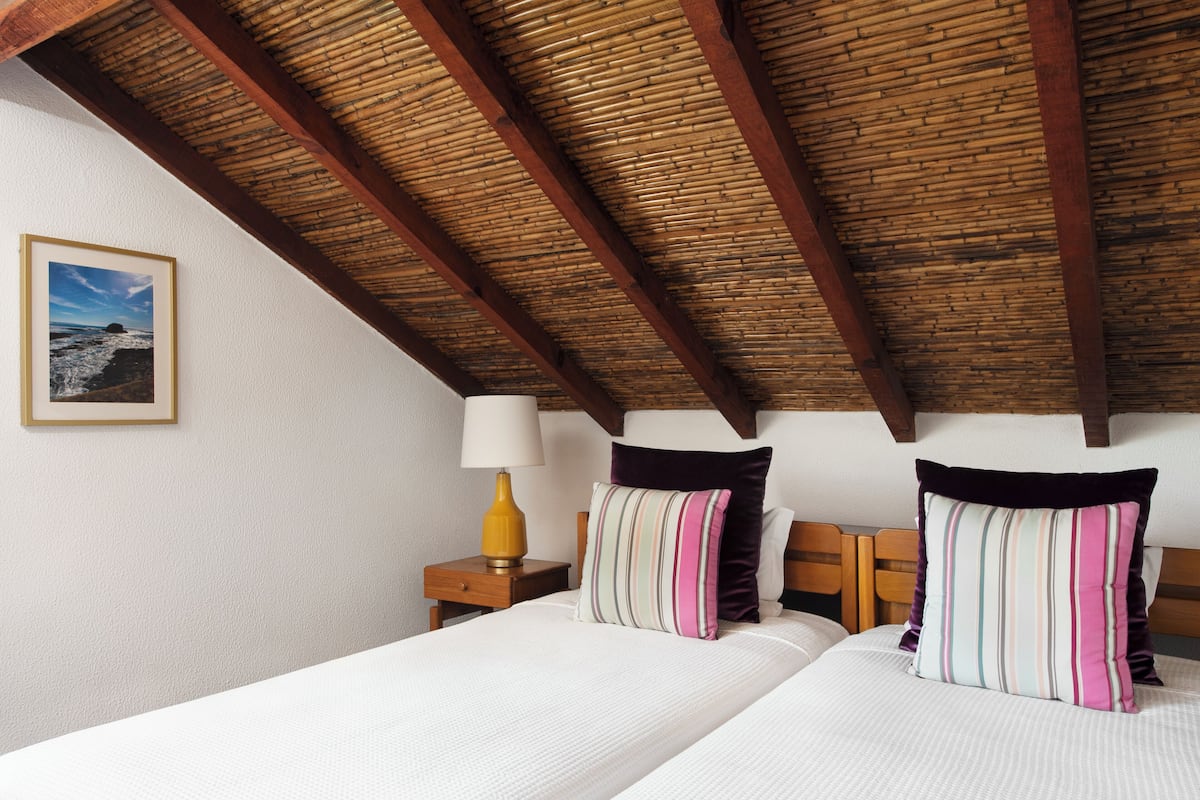 A bedroom with a wooden ceiling, two beds with striped pillows, a nightstand with a lamp, and a framed picture on the wall.
