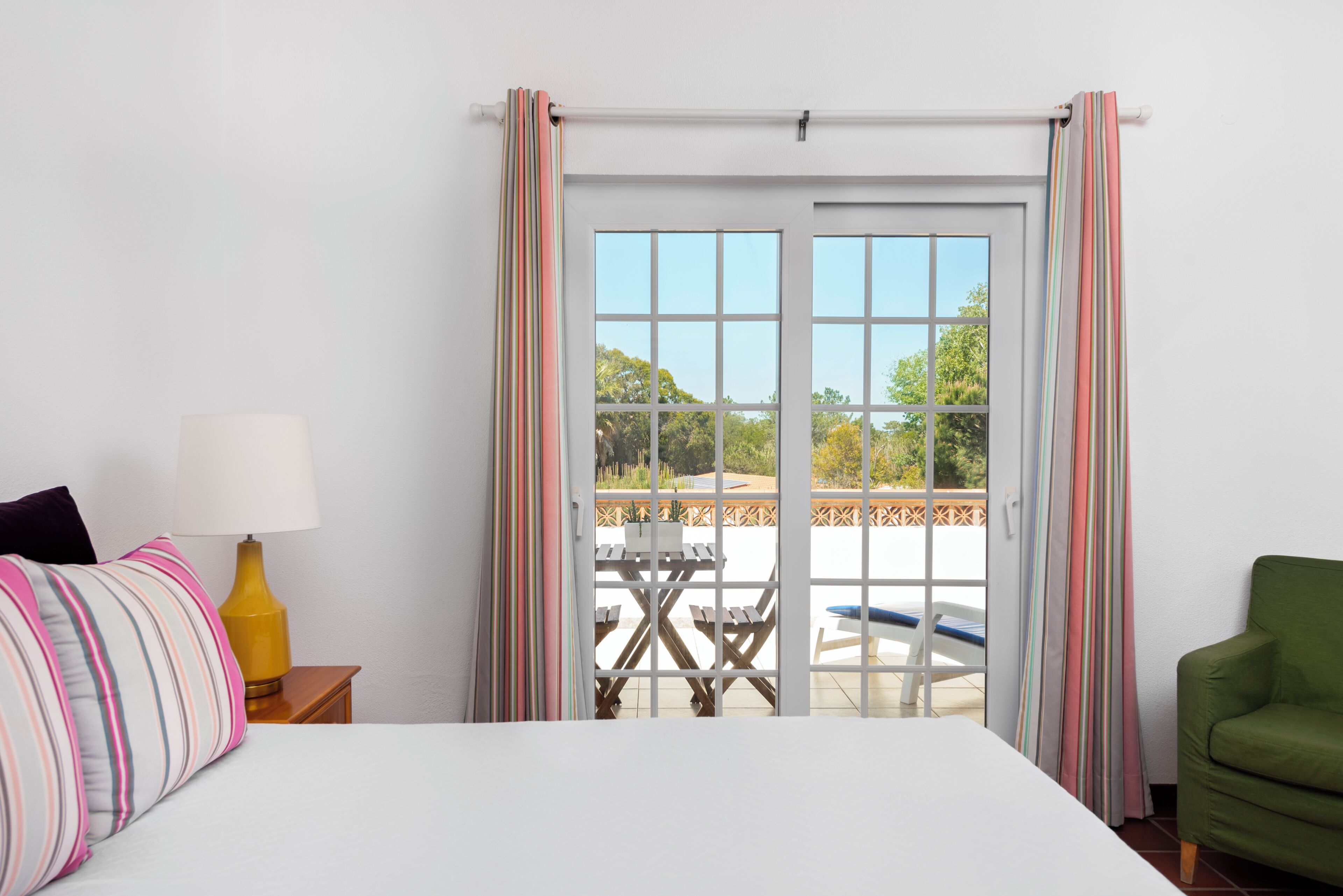 Une chambre d’hôtel avec un lit, une table de chevet avec une lampe, un fauteuil vert et une vue sur un patio extérieur avec une table et des chaises.
