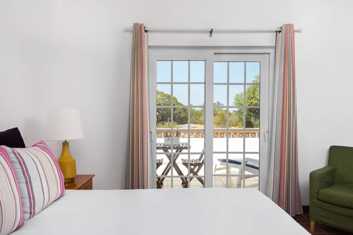 A hotel room with a bed, a nightstand with a lamp, a green armchair, and a view of an outdoor patio with a table and chairs.