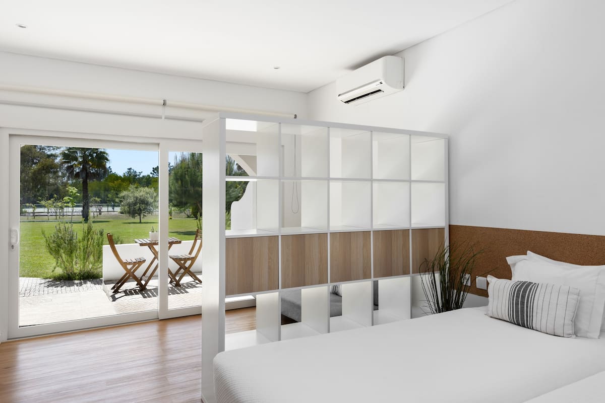 A modern bedroom with a large bed, a sliding glass door leading to an outdoor patio, and a view of a garden.