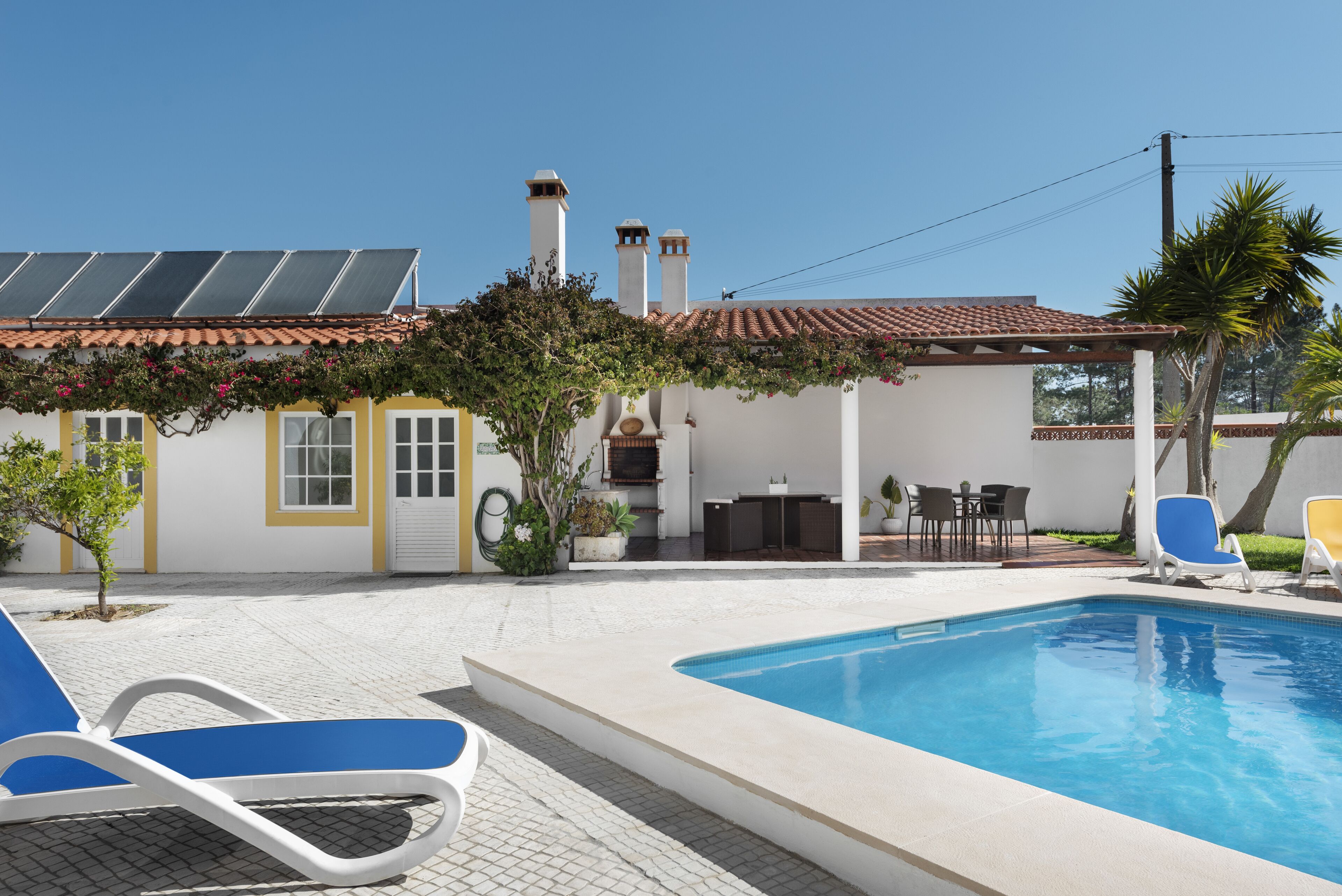 A swimming pool with lounge chairs, a patio with outdoor furniture, and a building with solar panels and chimneys.