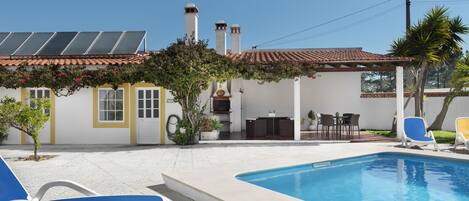 A swimming pool with lounge chairs, a patio with outdoor furniture, and a building with solar panels and chimneys.