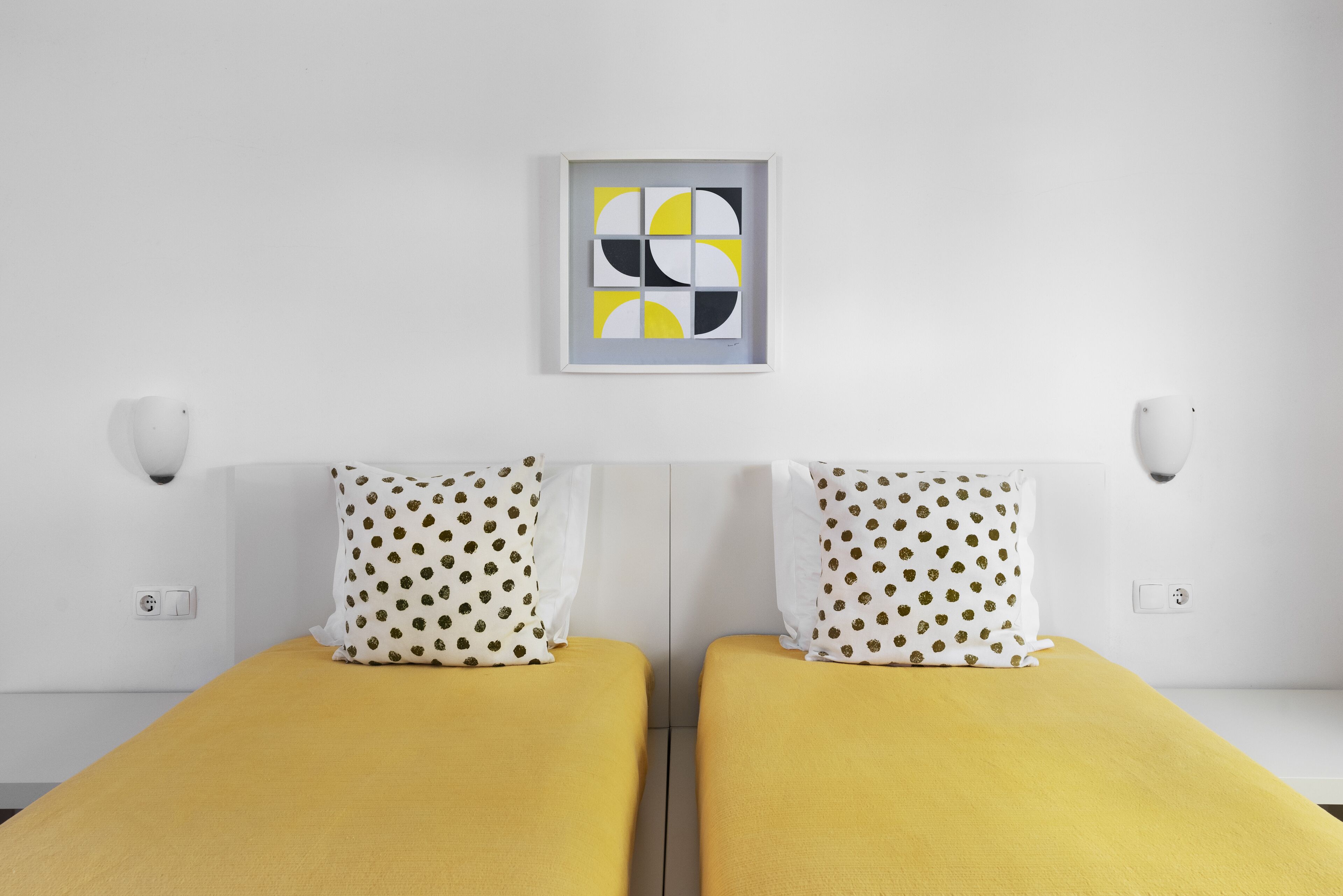 Habitación de hotel con dos camas, cada una con cubrecama amarillo y almohadas blancas con estampado marrón. Encima de las camas hay un cuadro abstracto enmarcado en la pared.