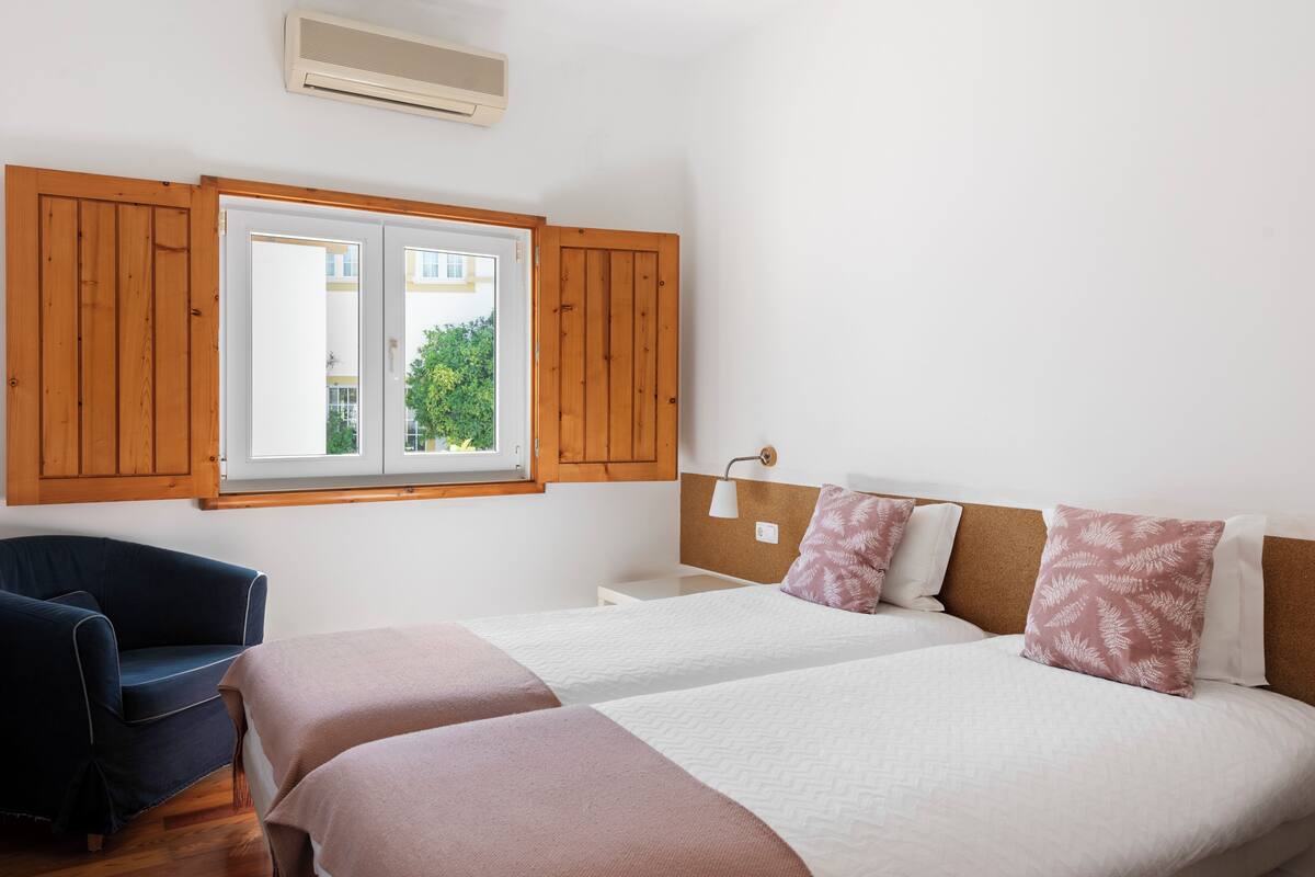 A bedroom with two beds, a wooden wardrobe, a chair, and a window with a view of greenery.