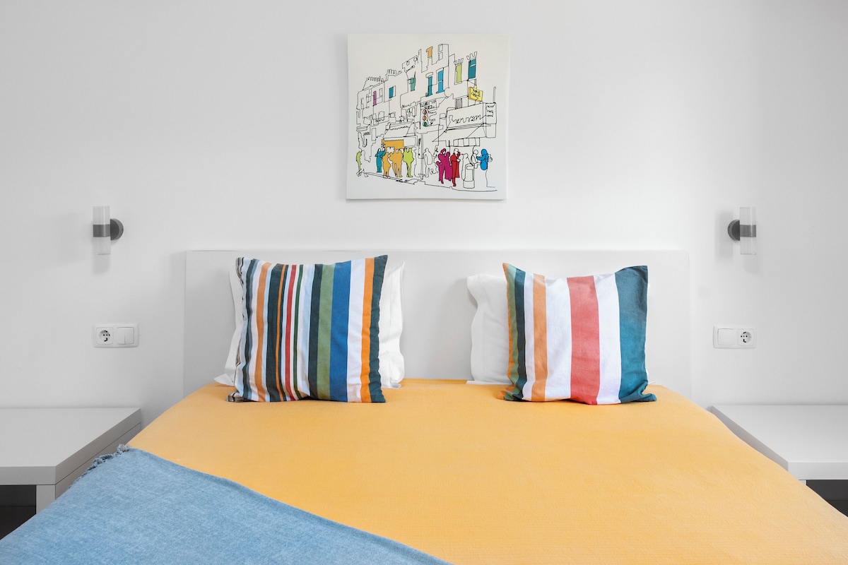 A bed with a yellow mattress, two striped pillows, and a wall-mounted artwork depicting a street scene.