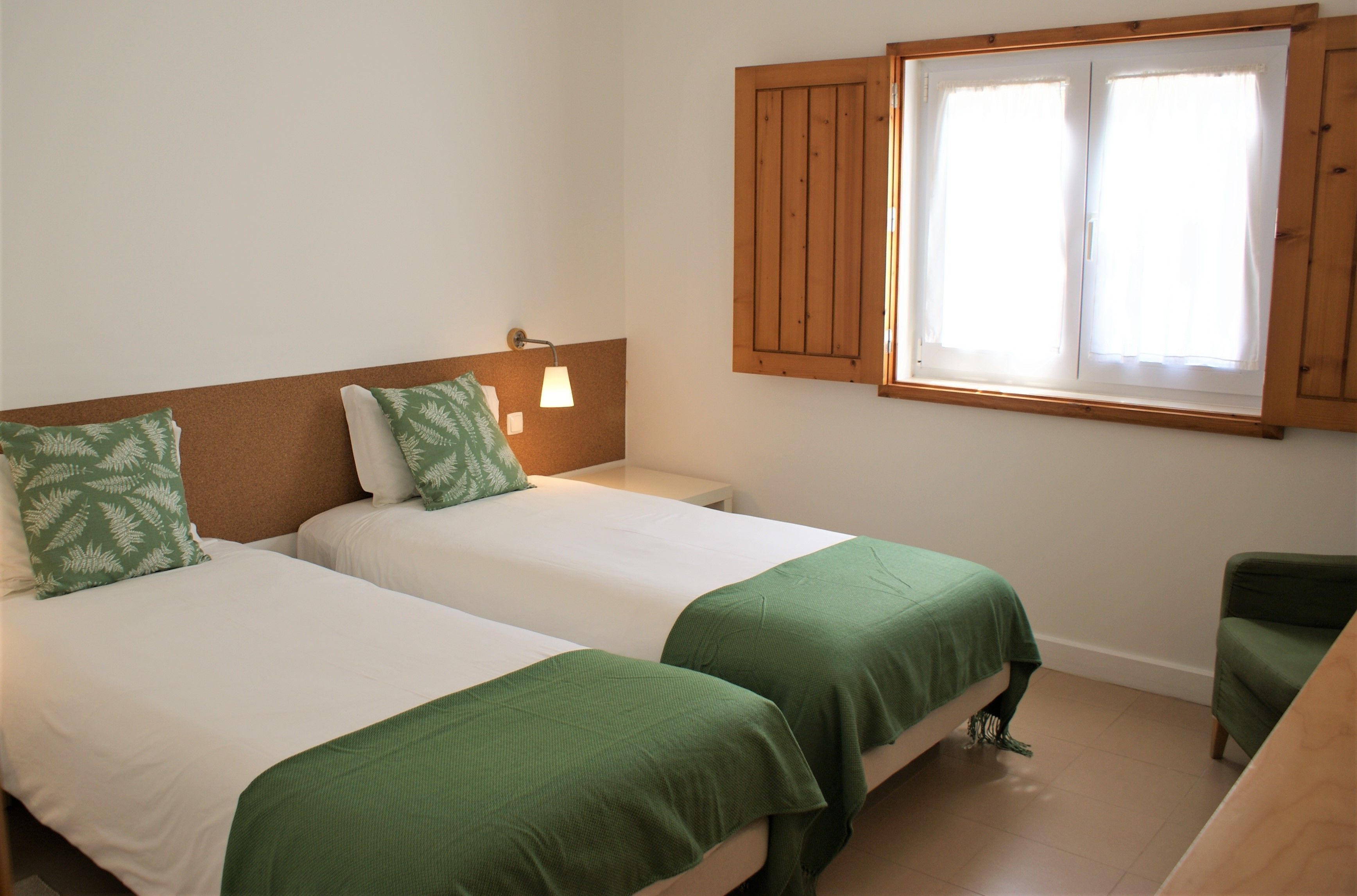 Habitación con dos camas, cada una con ropa de cama y almohadas verdes, una mesita de noche con una lámpara y una ventana con contraventanas de madera.