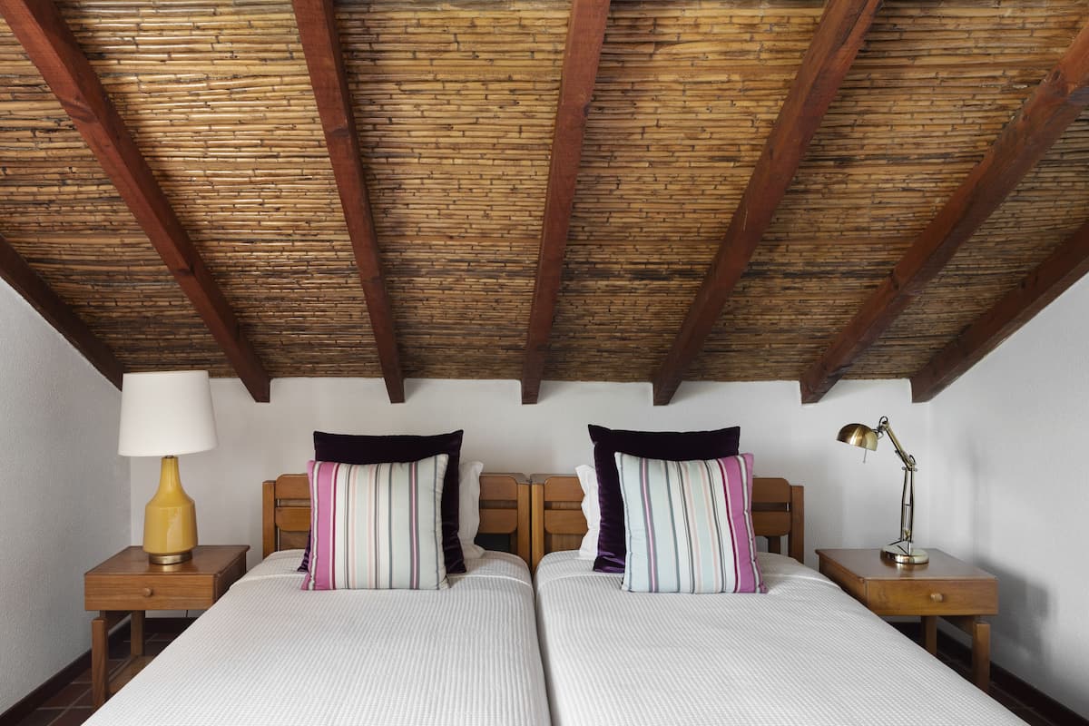 A room with two beds, wooden headboards, a wooden ceiling, a nightstand with a lamp, and a bedside table with a lamp.
