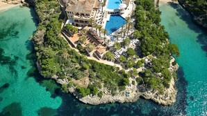Vista aérea de un complejo turístico costero con una amplia piscina, varias villas y abundante vegetación.