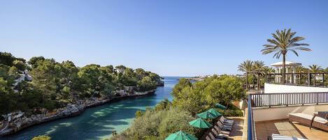 Un complejo turístico frente al mar con una terraza con vistas a un mar azul cristalino, vegetación exuberante y una palmera.