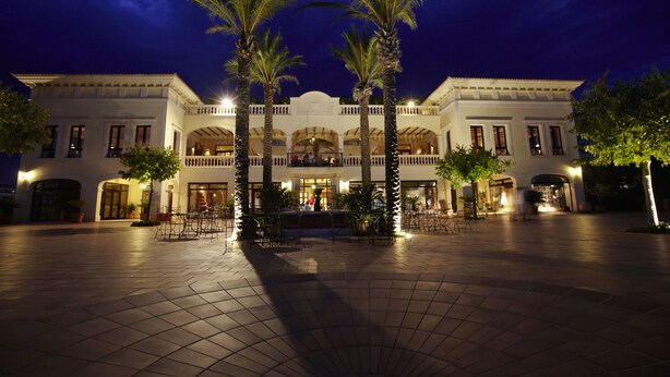 Un edificio grande y elegante con un patio central, palmeras y una entrada bien iluminada.