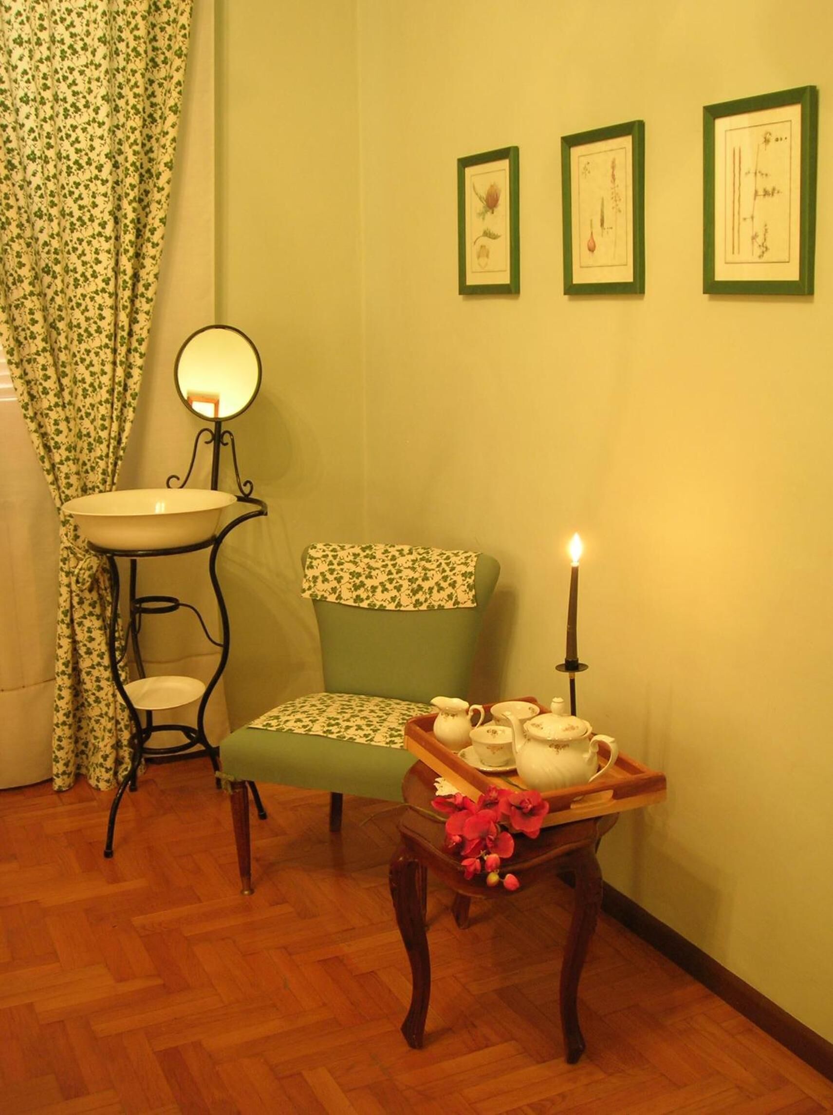 Ein gemütliches Zimmer mit einem grünen Sessel, einem kleinen runden Tisch mit einer Teeservice und einem Wandspiegel, sowie gerahmten botanischen Drucken.