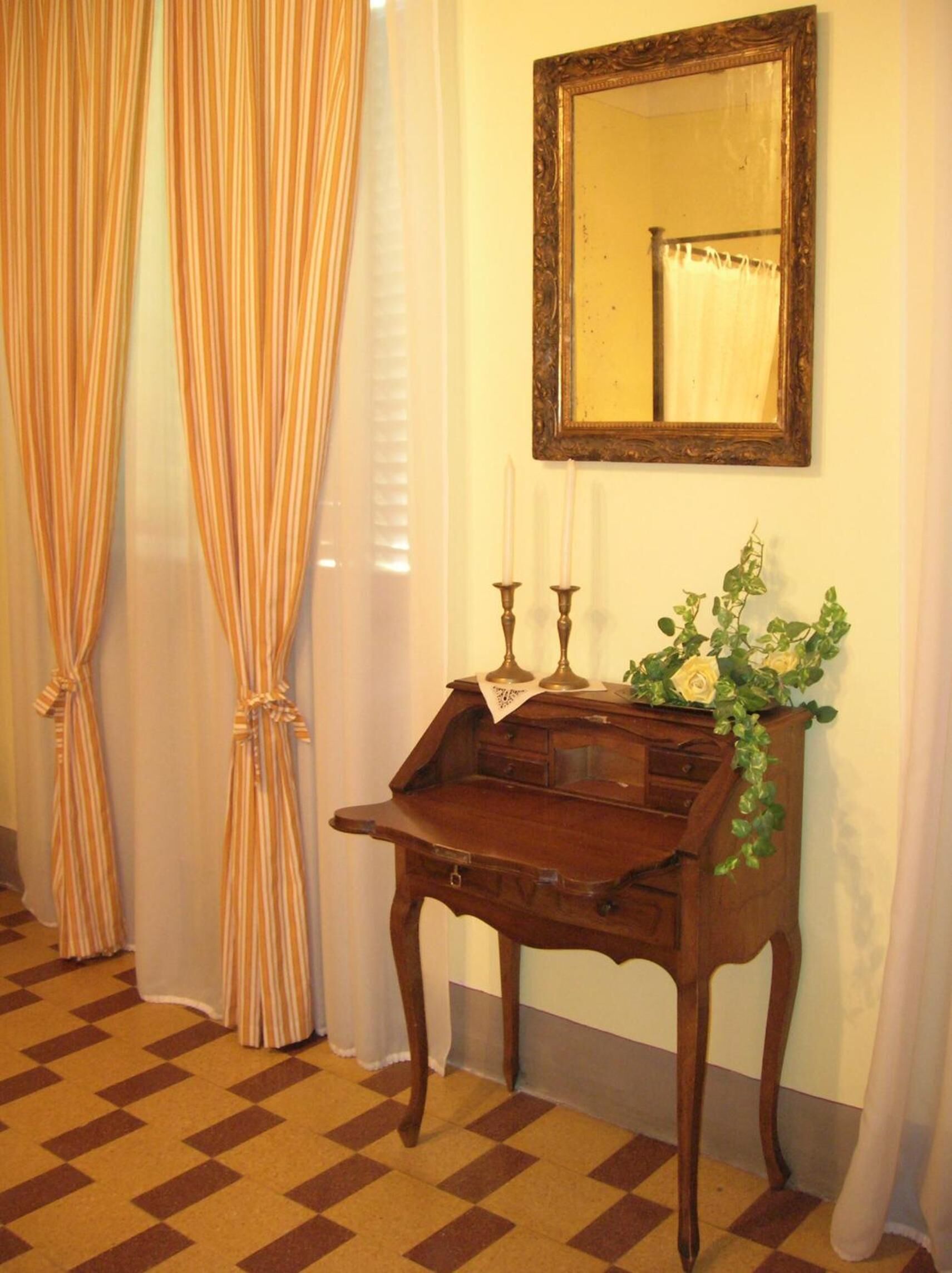 Une pièce avec un sol à carreaux, un bureau en bois doté de tiroirs, un miroir au mur et un vase rempli de fleurs.