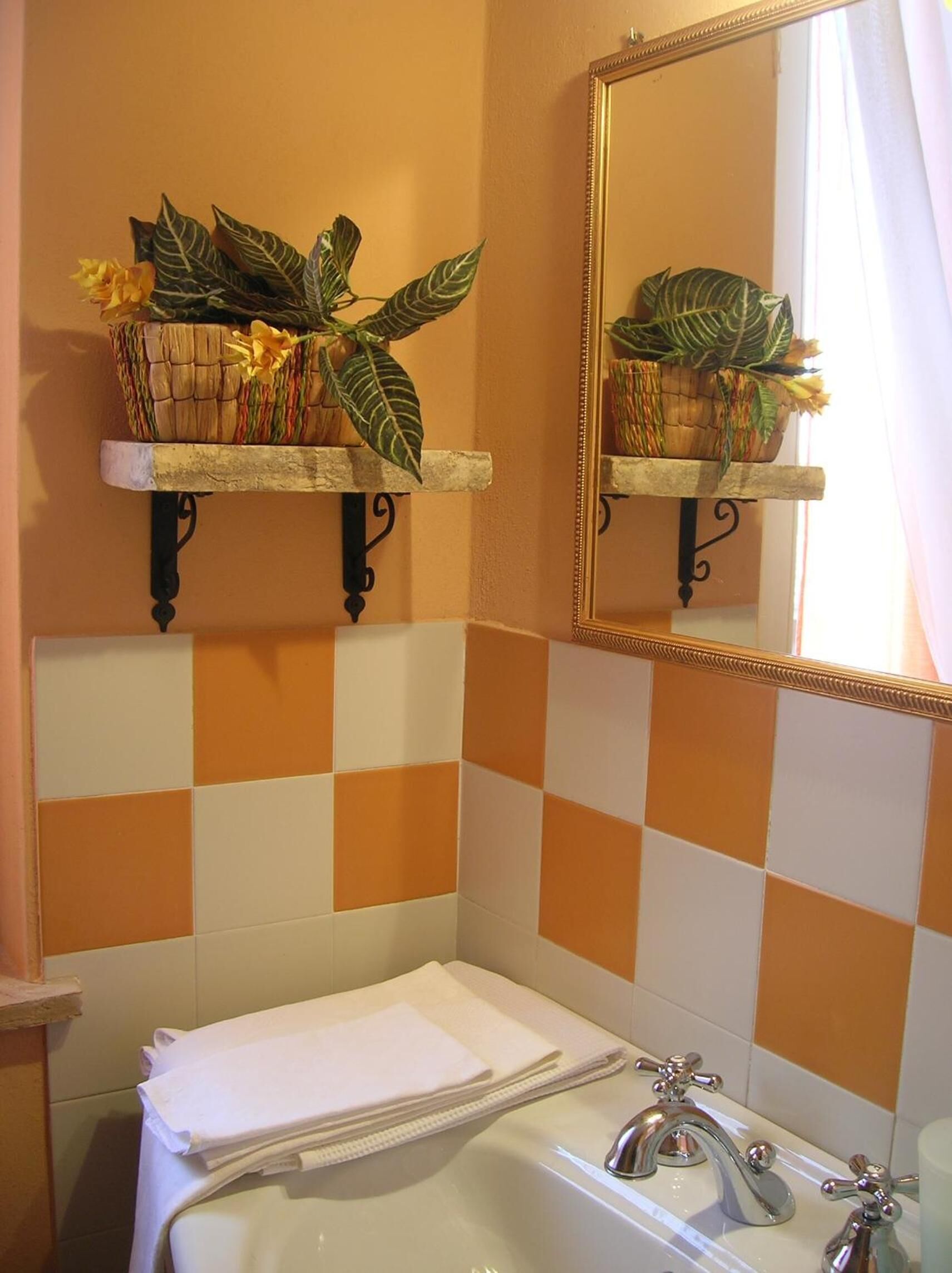 A bathroom with orange and white checkered tiles, a white bathtub, two wall-mounted shelves with potted plants, and a mirror with a gold frame.