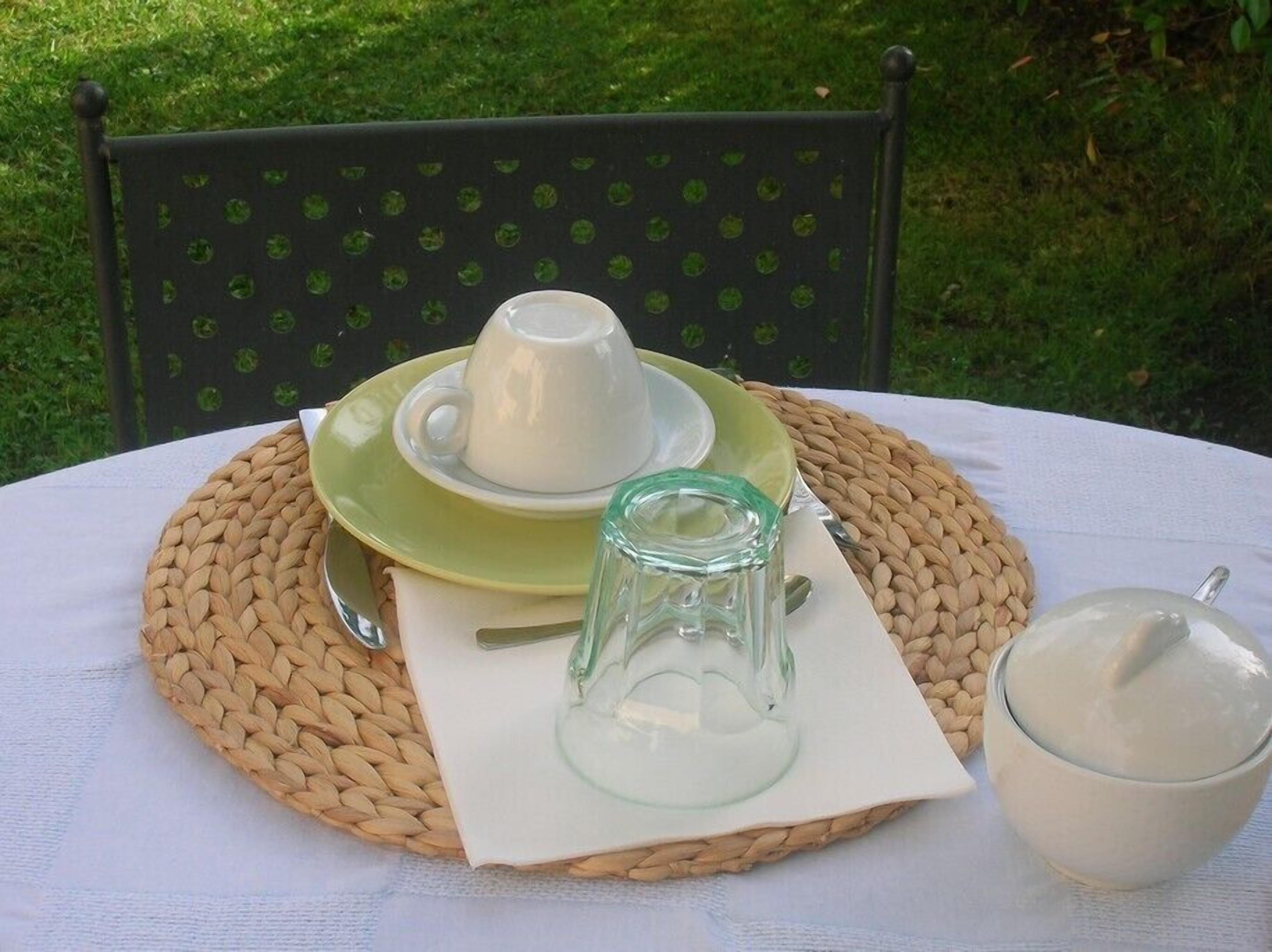 A table set outdoors with a woven placemat, a white cup and saucer, a glass pitcher, and a bowl of almonds.