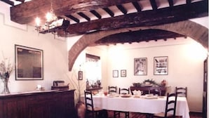 Un comedor con una mesa larga dispuesta para cuatro personas, vigas de madera en el techo y cuadros en las paredes.