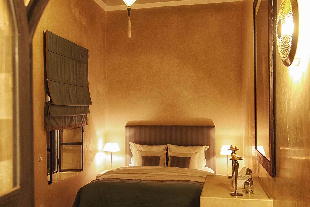 A hotel room with a bed, a chair, a bedside table, a mirror, and a window with curtains.