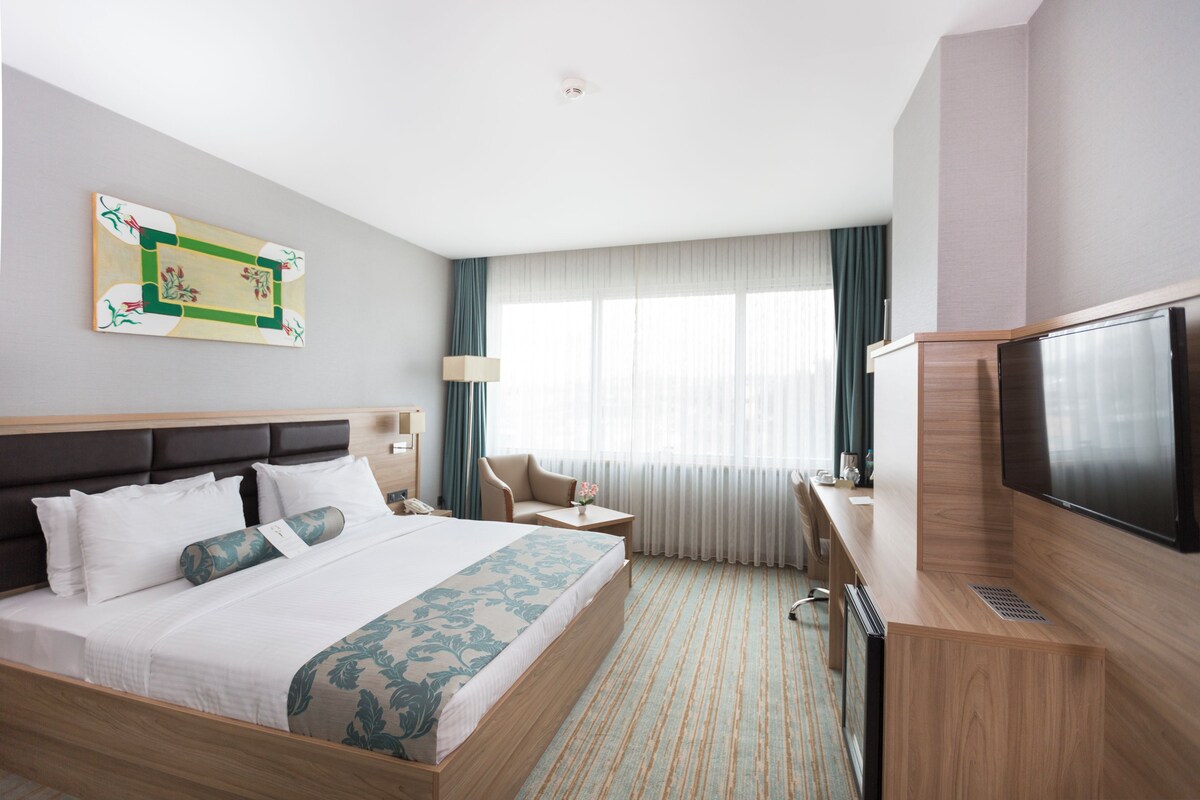 A hotel room with a large bed, a desk with a flat-screen TV, a chair, a lamp, and a window with curtains.