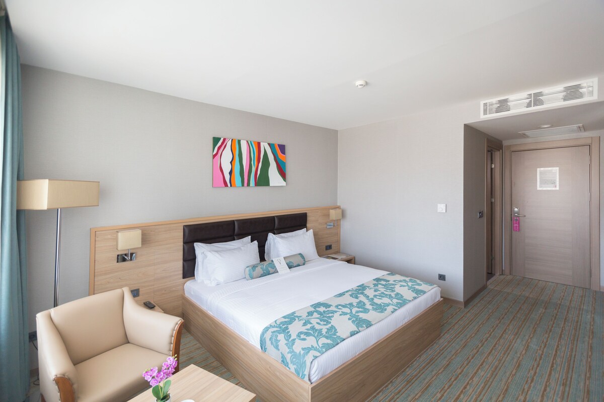 Habitación de hotel con cama, una silla, una mesita de noche, lámpara y una obra de arte en la pared.