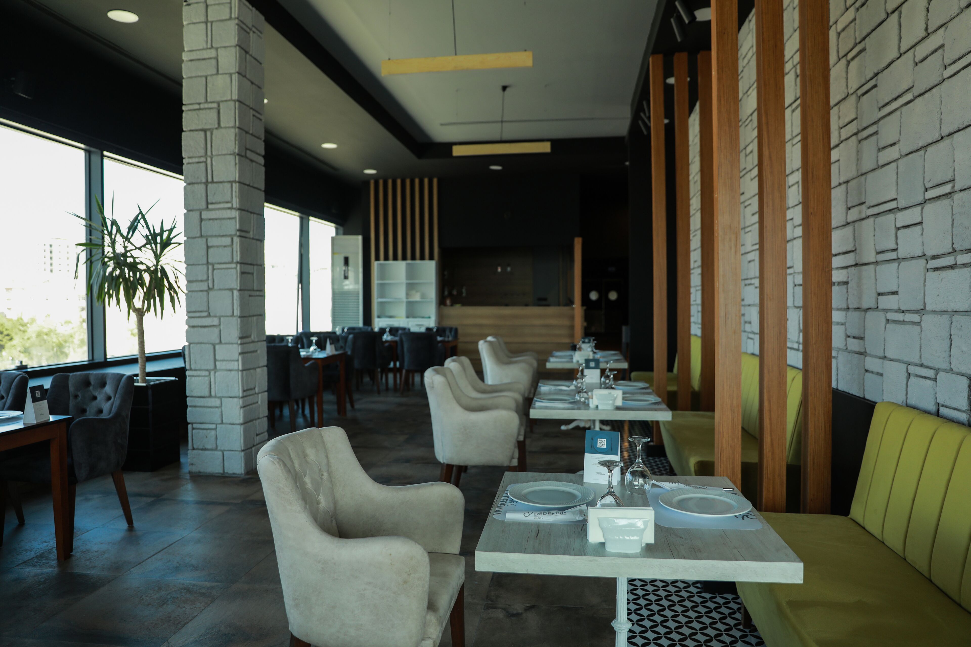 Ein modernes Restaurant mit hohen Tischen, gepolsterten Stühlen, großen Fenstern und einer Wandgestaltung aus Backstein.