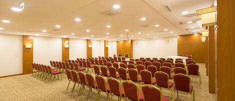A conference room with rows of red chairs and tables, carpeted floor, and recessed ceiling lights.