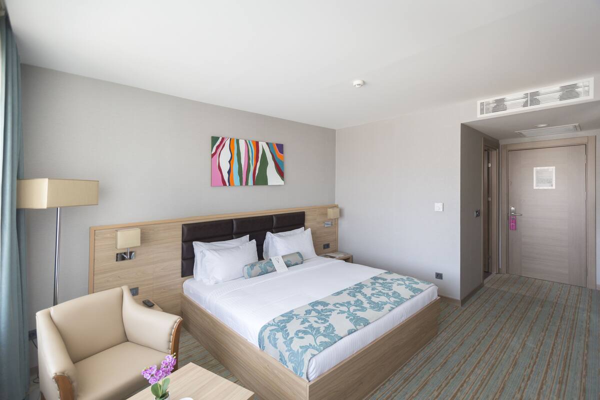 Habitación de hotel con cama, una silla, una mesita de noche, lámpara y una obra de arte en la pared.