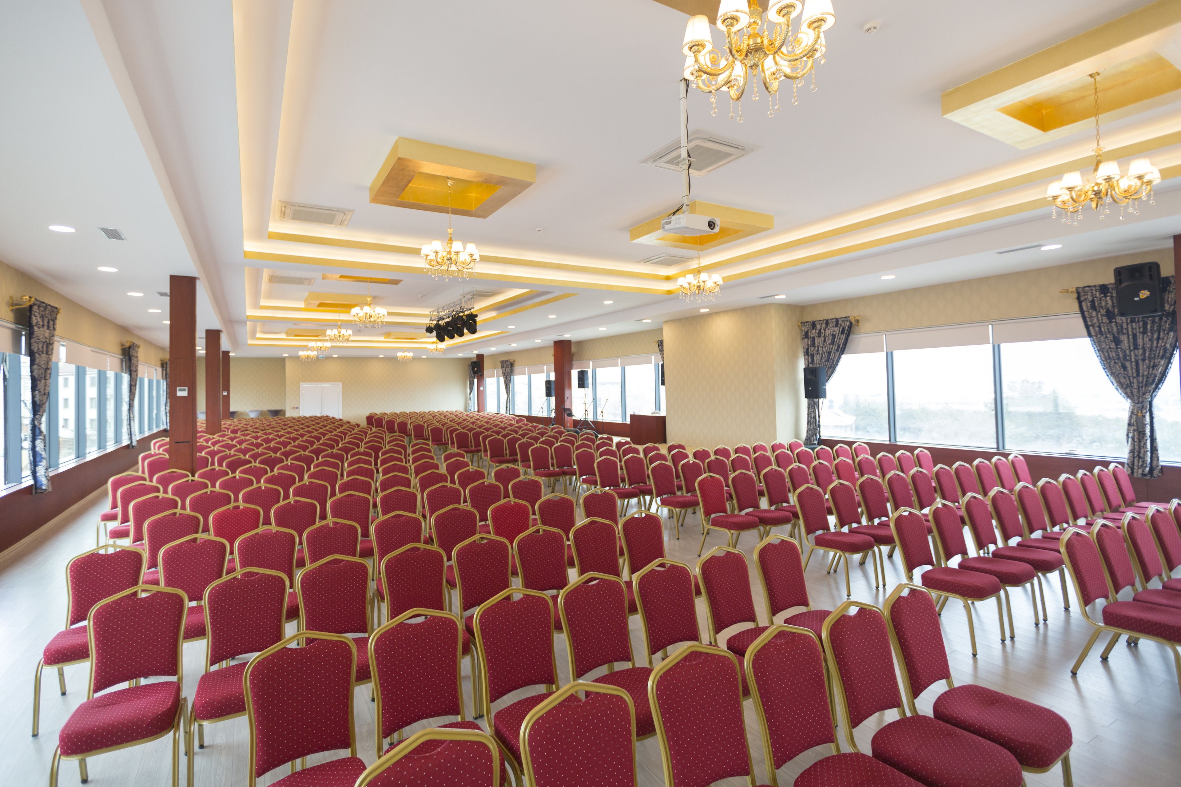 A large hall with rows of red chairs and gold-framed chairs, chandeliers, and large windows.