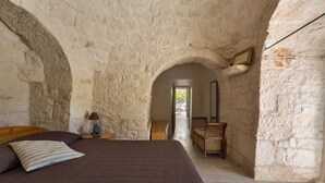 A bedroom with a stone wall, a bed with a brown quilt, a nightstand with a lamp, and a wooden bench. - Trullo Luna (Ostuni)